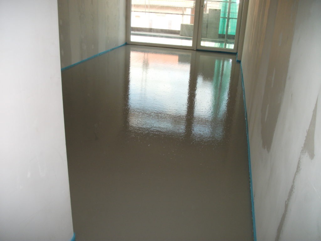 Świeżo wylana, samopoziomująca posadzka z widocznym odbiciem okna i zabezpieczającą, niebieską taśmą przy ścianach.