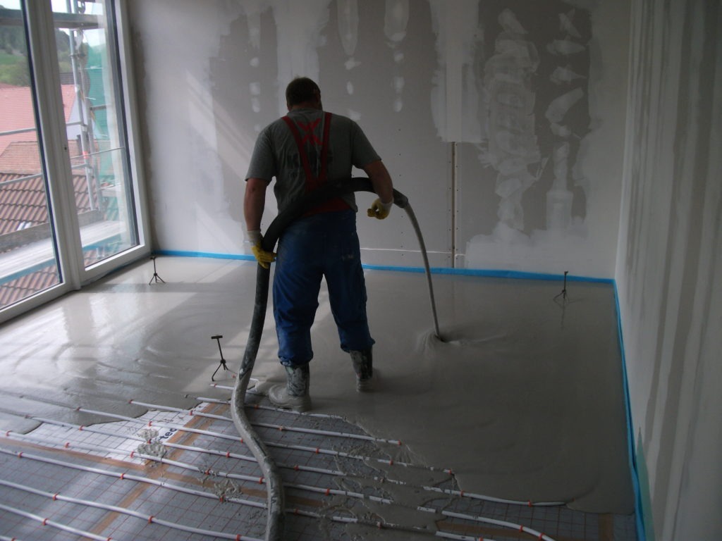 Aplikacja wylewki samopoziomującej na ogrzewanie podłogowe w jasnym pomieszczeniu z dużym oknem; widoczny pracownik w gumowych butach rozprowadzający masę za pomocą węża.