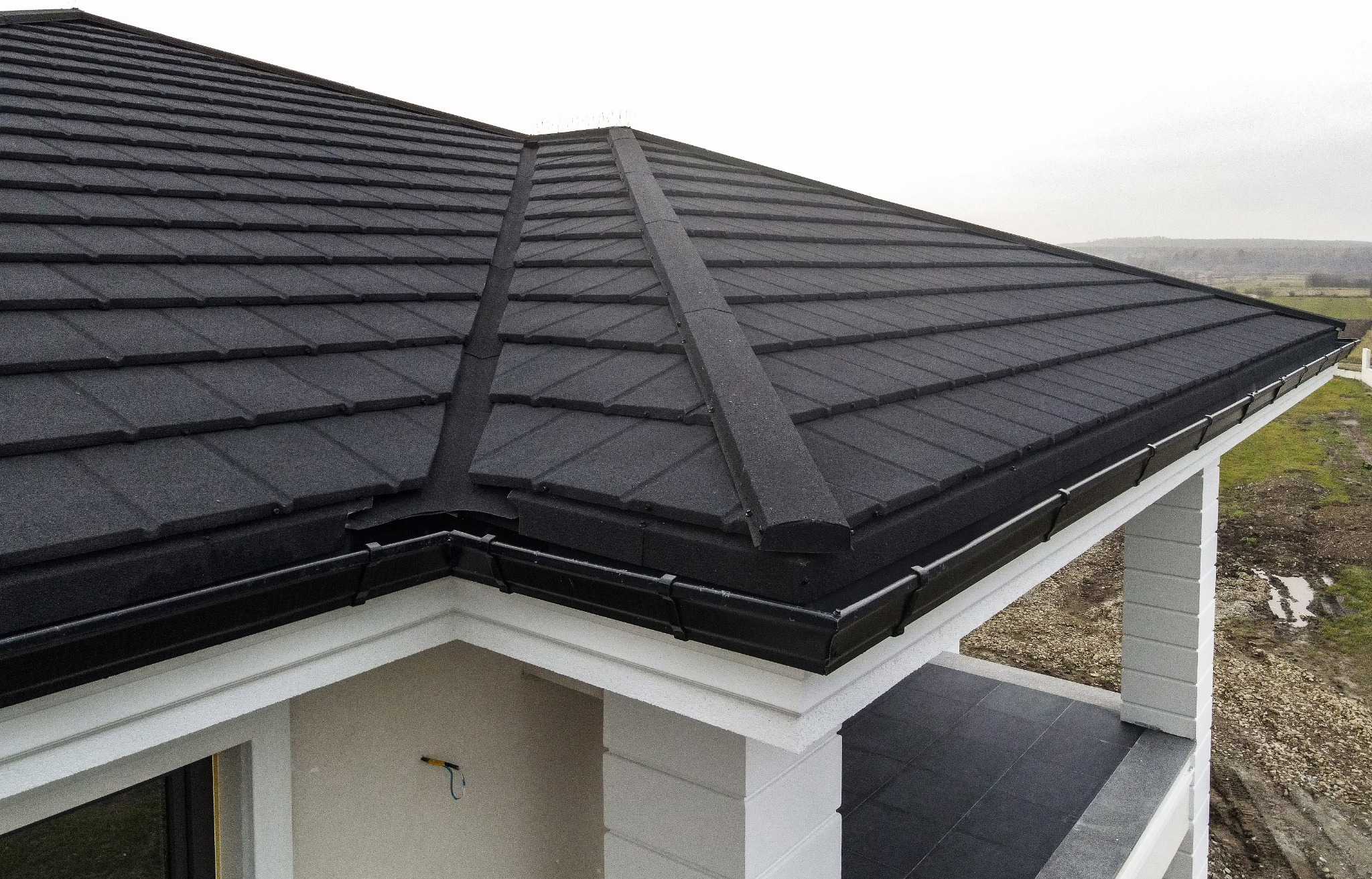Nowy, grafitowy dach z blachodachówki modułowej, z zamontowanymi czarnymi rynnami, widok z góry na narożnik budynku.