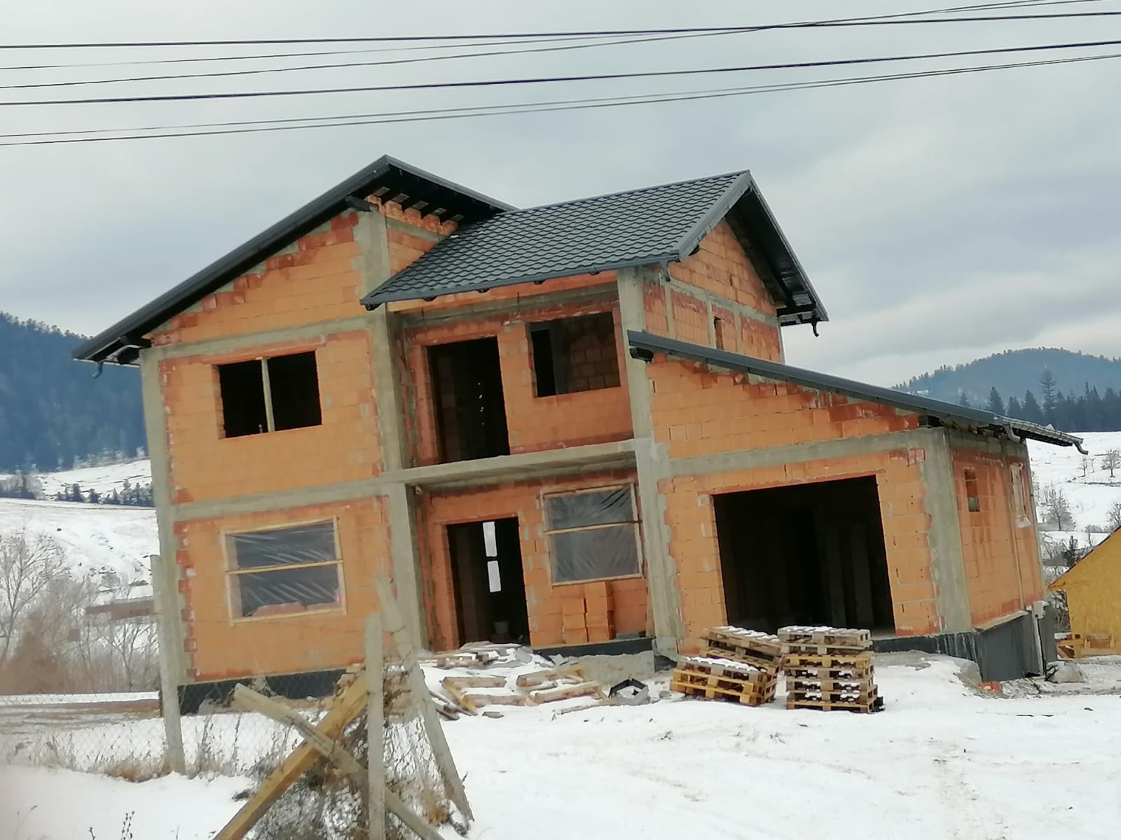 Surowy stan dwukondygnacyjnego domu z czerwonej cegły z zamontowanym ciemnoszarym dachem, widoczne góry w tle i śnieg na pierwszym planie.
