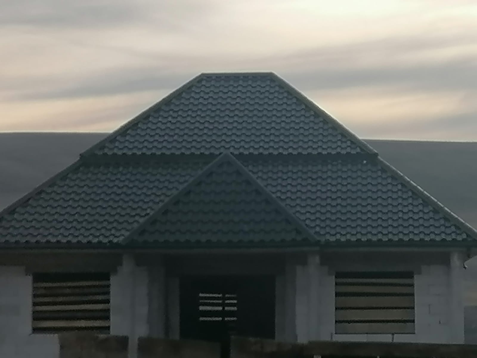 Dom w stanie surowym z nowym, ciemnozielonym dachem z blachodachówki trapezowej i zamontowanymi rynnami, widok z dołu na tle pochmurnego nieba.