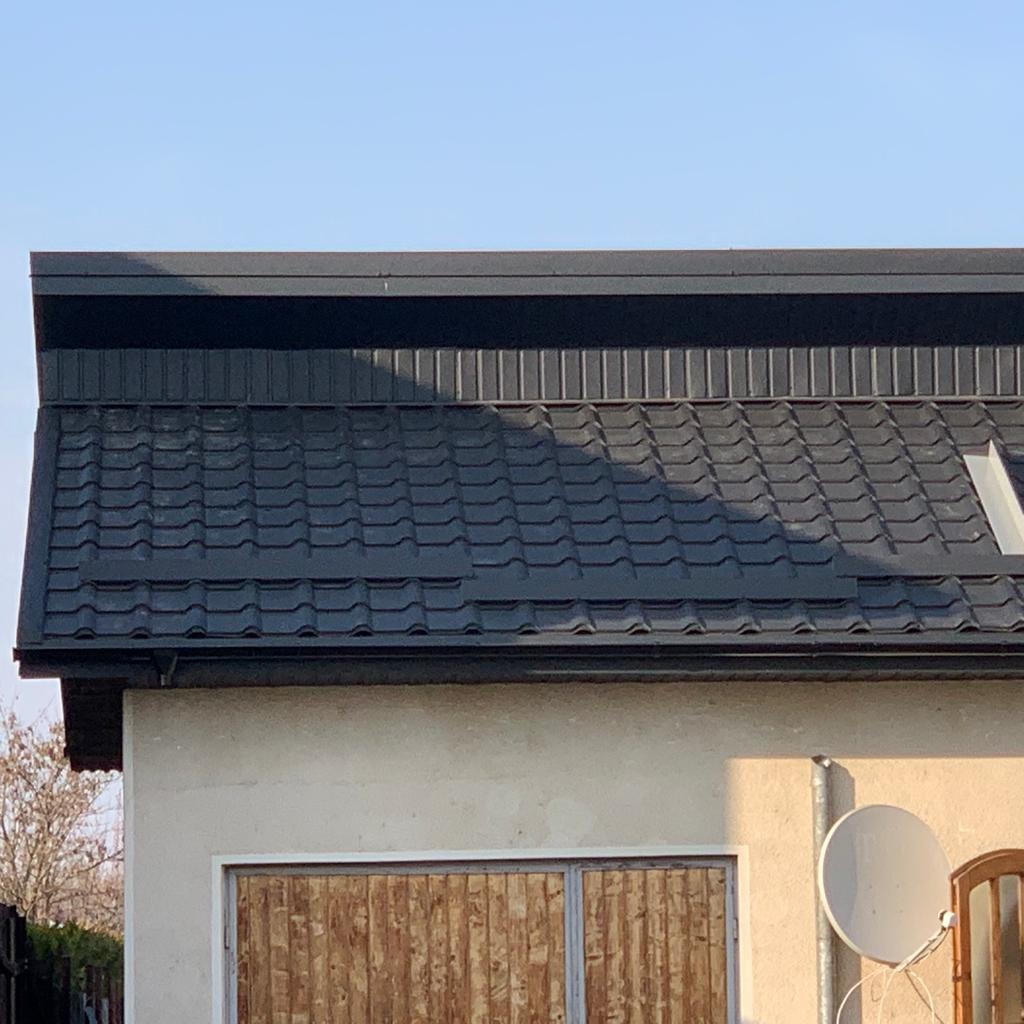 Ciemny dach z metalowej dachówki modułowej, widoczny fragment elewacji budynku z drewnianymi wrotami garażowymi i anteną satelitarną.