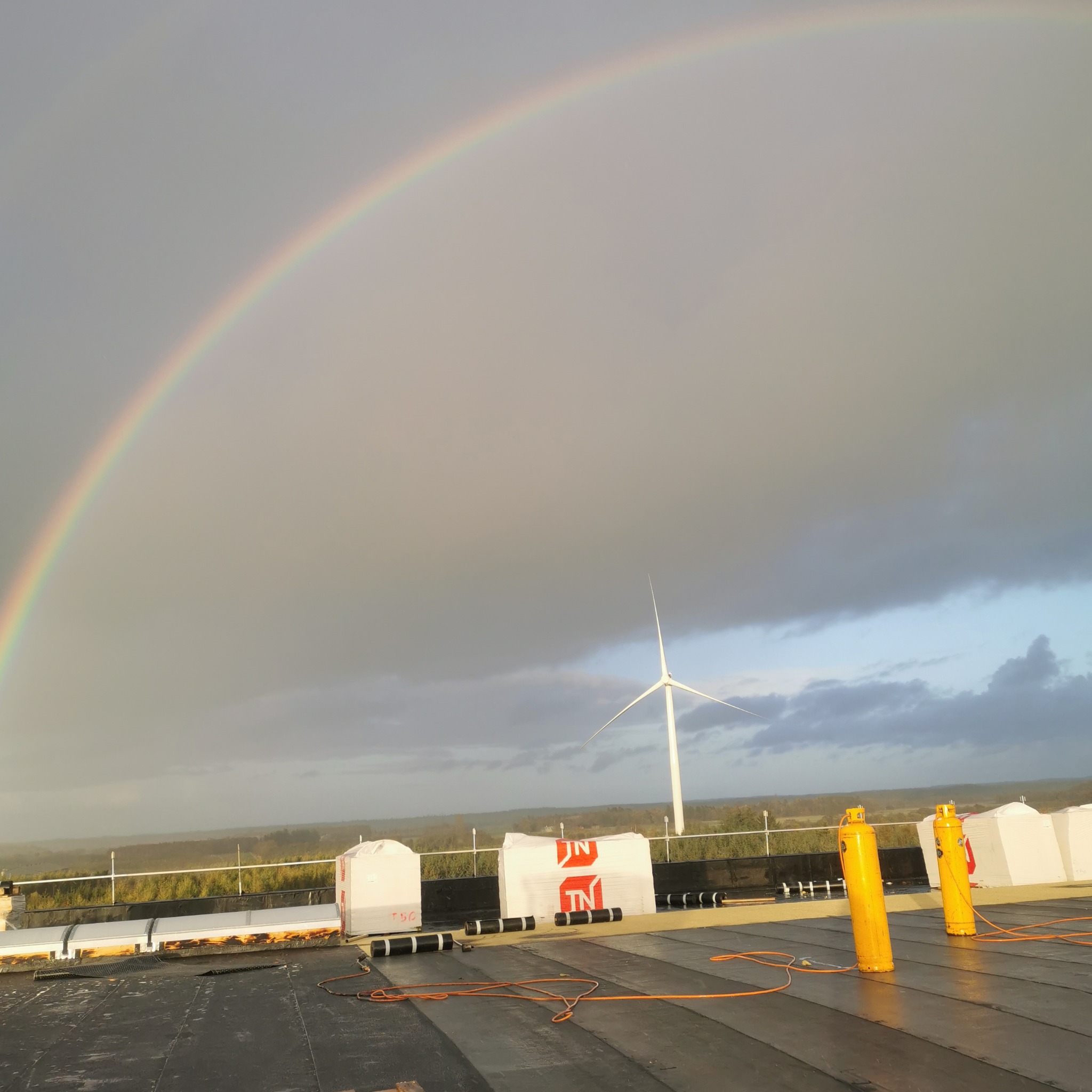 Widok na czarny, płaski dach z rolkami papy, butlami gazowymi i paczkami materiałów budowlanych na tle krajobrazu z turbiną wiatrową i tęczą na pochmurnym niebie.