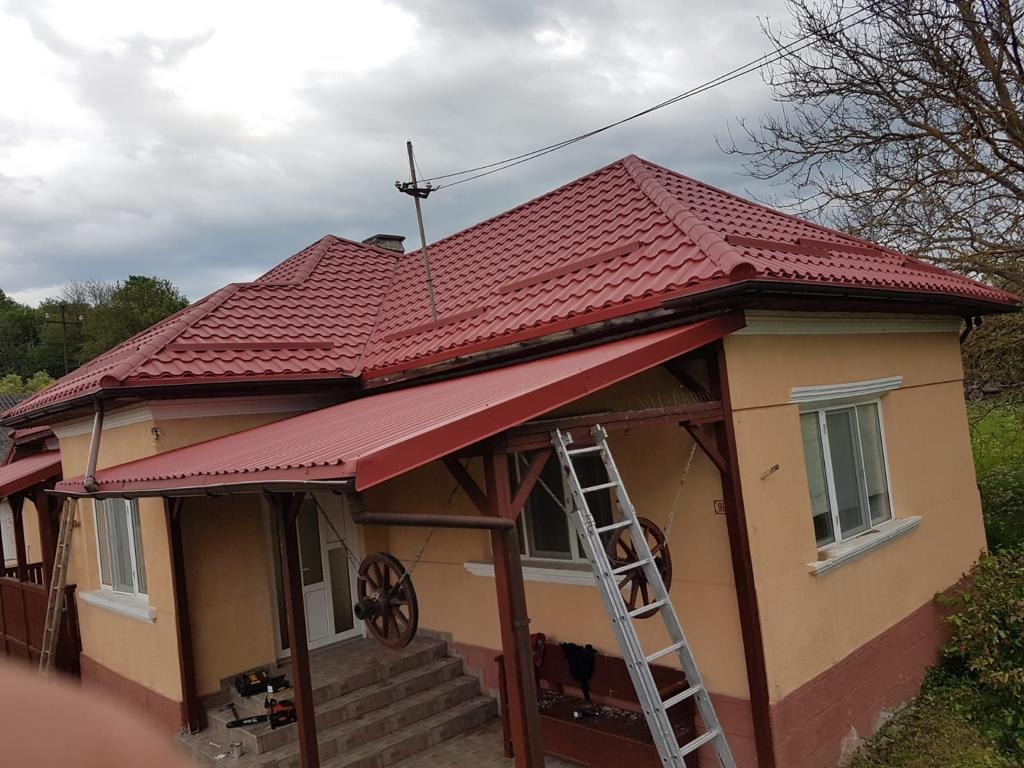 Dom z nowym, czerwonym dachem z blachodachówki, widoczna drabina oparta o ścianę, teren wokół domu częściowo uporządkowany, pochmurne niebo.
