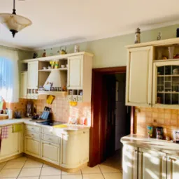 Jasna kuchnia z kremowymi szafkami w stylu rustykalnym, z dekoracyjnymi przedmiotami na półkach i mozaiką na ścianie, oświetlona naturalnym światłem wpadającym przez okno.