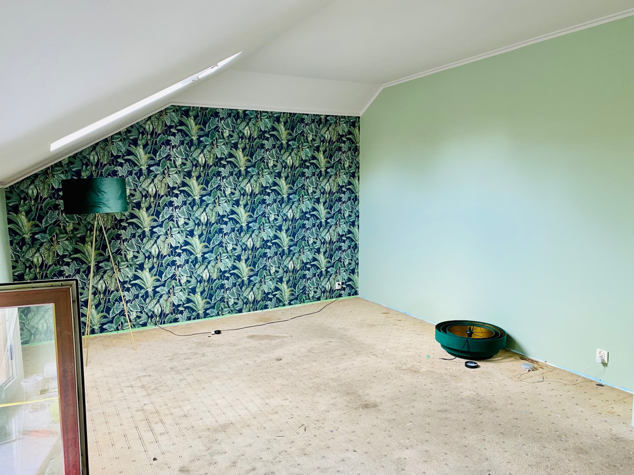 Pokój na poddaszu po częściowym remoncie: jedna ściana z tapetą w tropikalne liście, druga pomalowana na zielono, stara wykładzina dywanowa, widoczna lampa podłogowa i elementy oświetlenia sufitowego.