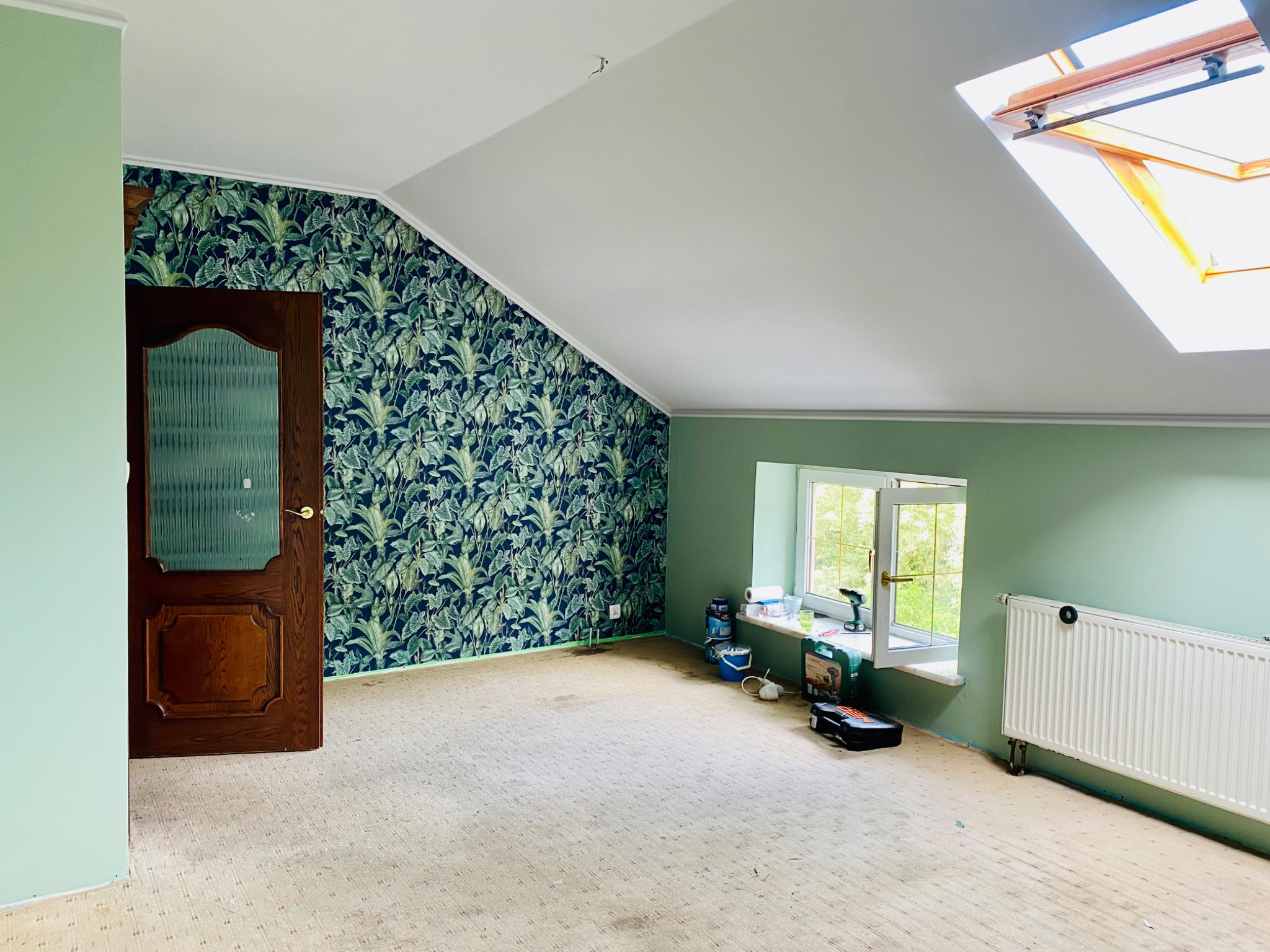 Poddasze z oknem dachowym i zielonymi ścianami, jedna ze ścian pokryta tapetą w tropikalne liście, widoczne narzędzia malarskie i grzejnik, przygotowane do dalszych prac.