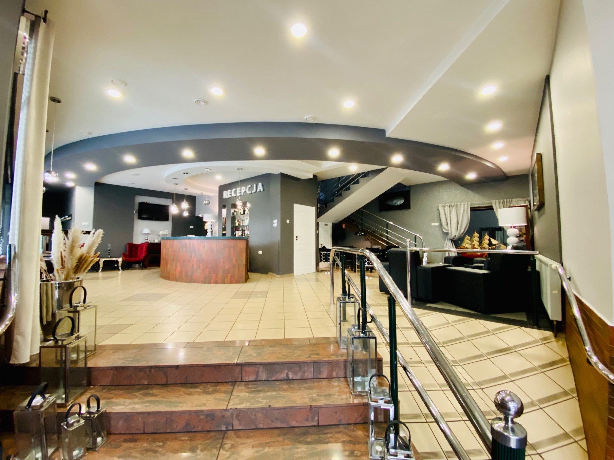 Eleganckie wnętrze hotelu z recepcją, schodami i dekoracyjnymi lampionami na stopniach, oświetlone punktowymi lampami sufitowymi.