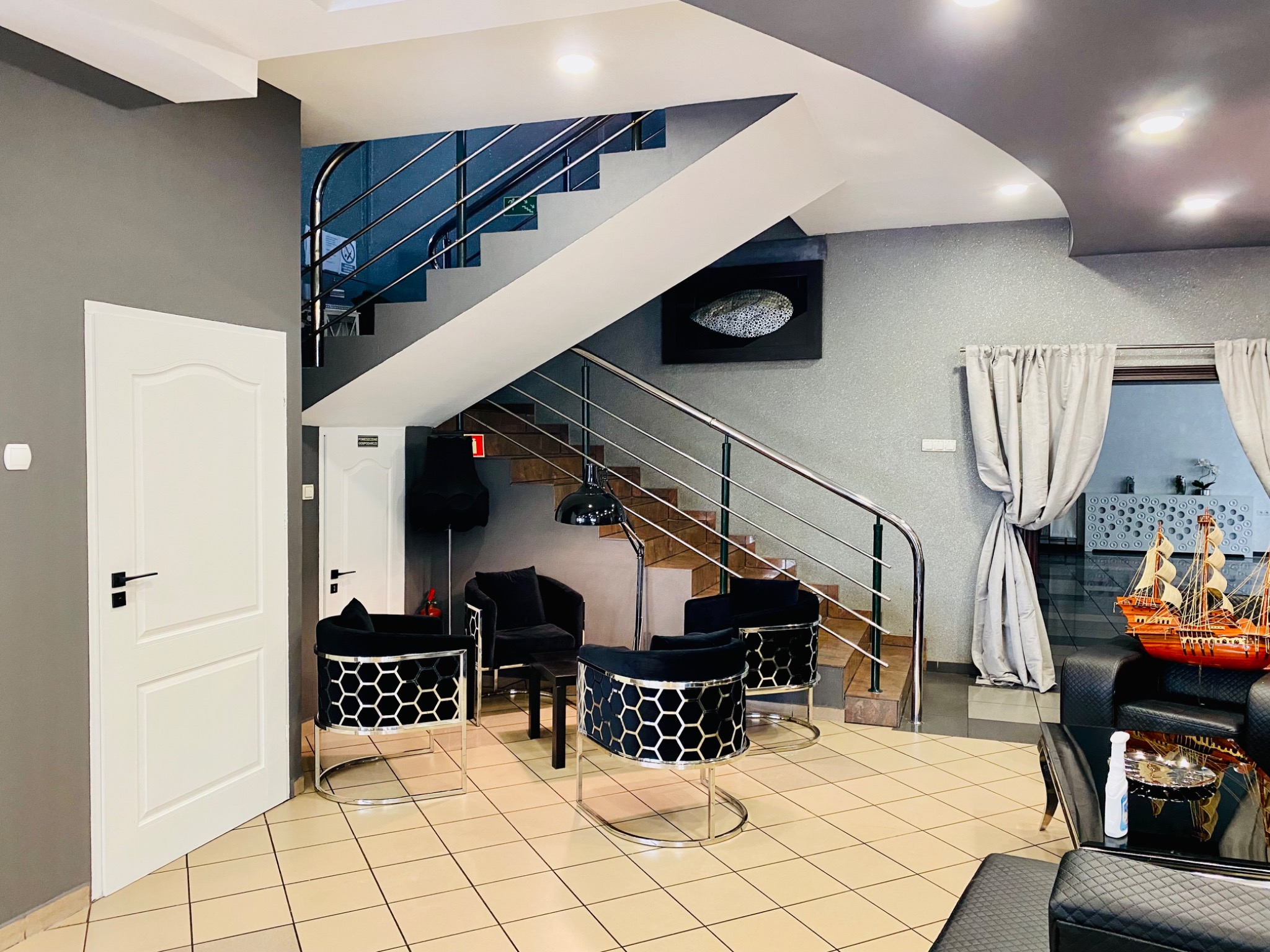 Nowoczesne wnętrze z szarymi ścianami, białymi drzwiami i schodami z metalową balustradą. W centralnej części cztery czarne krzesła z metalowymi elementami w kształcie plastra miodu. Na podłodze...