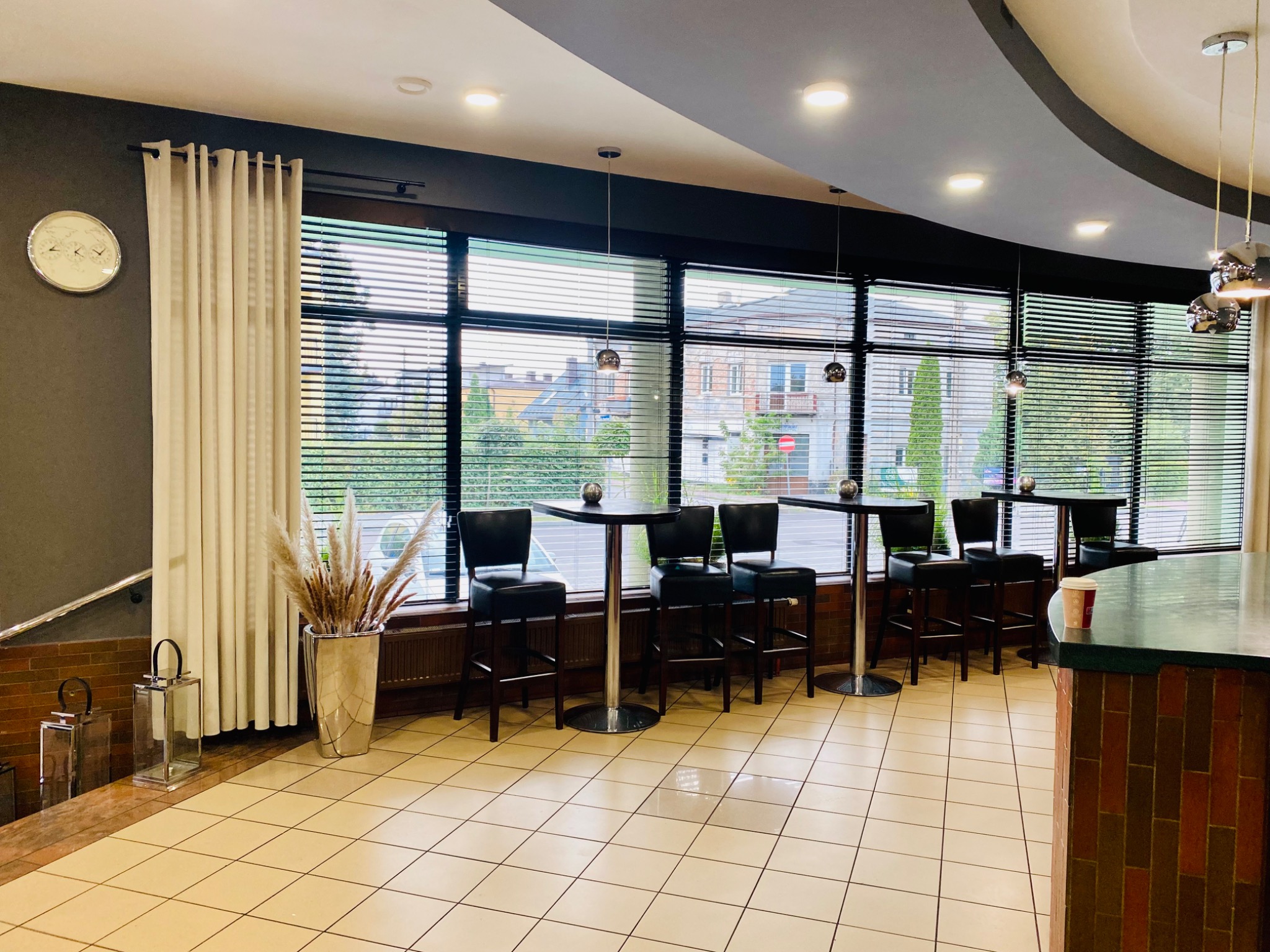 Wnętrze biura z wysokimi stolikami barowymi i krzesłami, widokiem na zewnątrz przez żaluzje, dekoracyjnymi lampami wiszącymi, oraz elementami dekoracyjnymi jak wazon z trawami i lampiony.