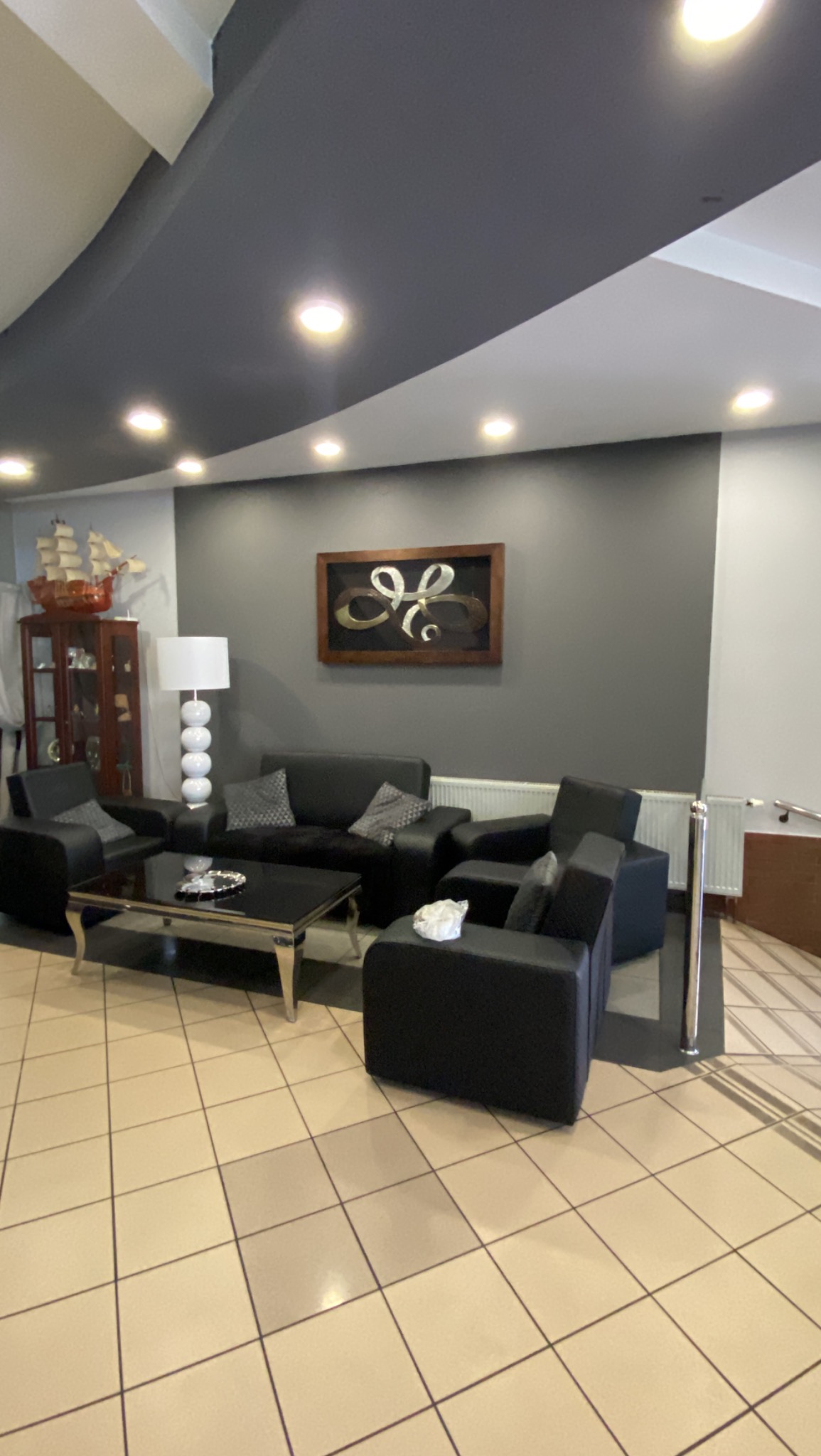 Ciemnoszara ściana z abstrakcyjną dekoracją w ramie nad kompletem wypoczynkowym z czarnej skóry; w tle witryna z modelem żaglowca i lampa z kulistą podstawą.