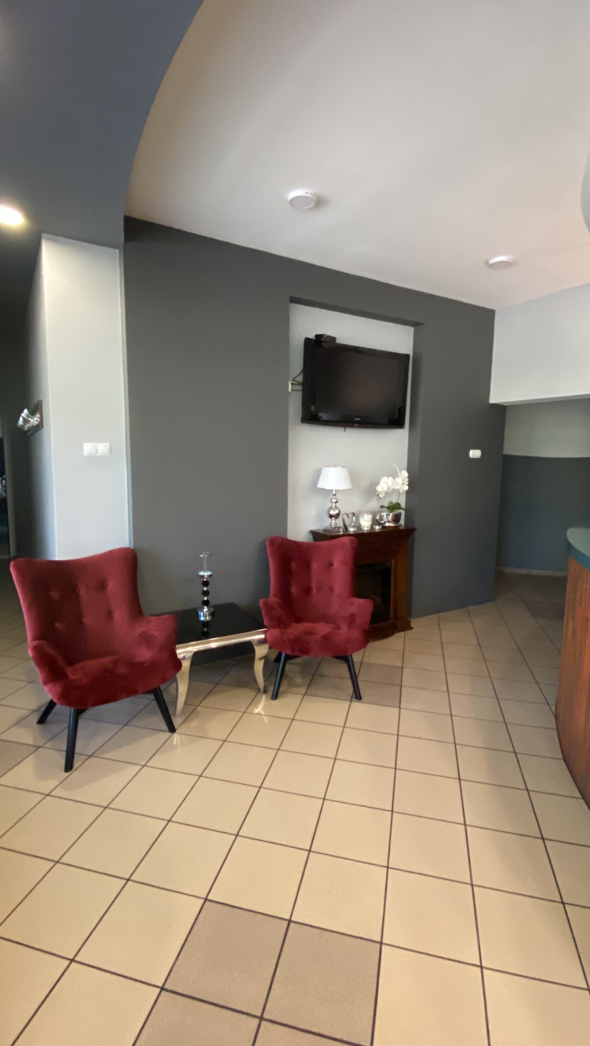 Nowoczesna aranżacja wnętrza biura z dwoma czerwonymi, welurowymi fotelami, czarnym stolikiem kawowym i telewizorem na szarej ścianie; widoczna lampka nocna i dekoracje.