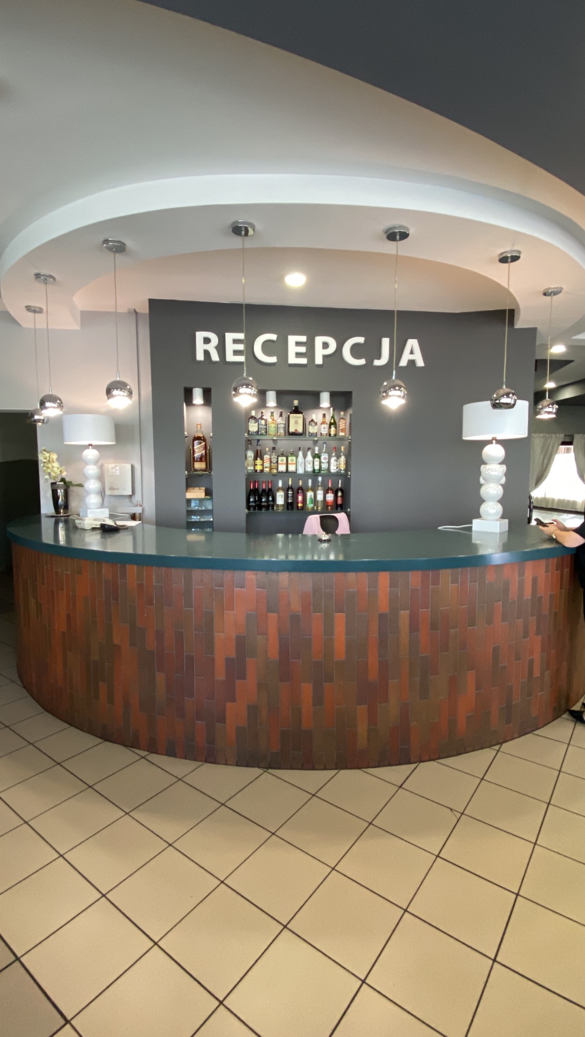 Recepcja z ladą wykończoną brązowymi płytkami, podświetlana półka z alkoholami, wiszące lampy i białe lampy stołowe.