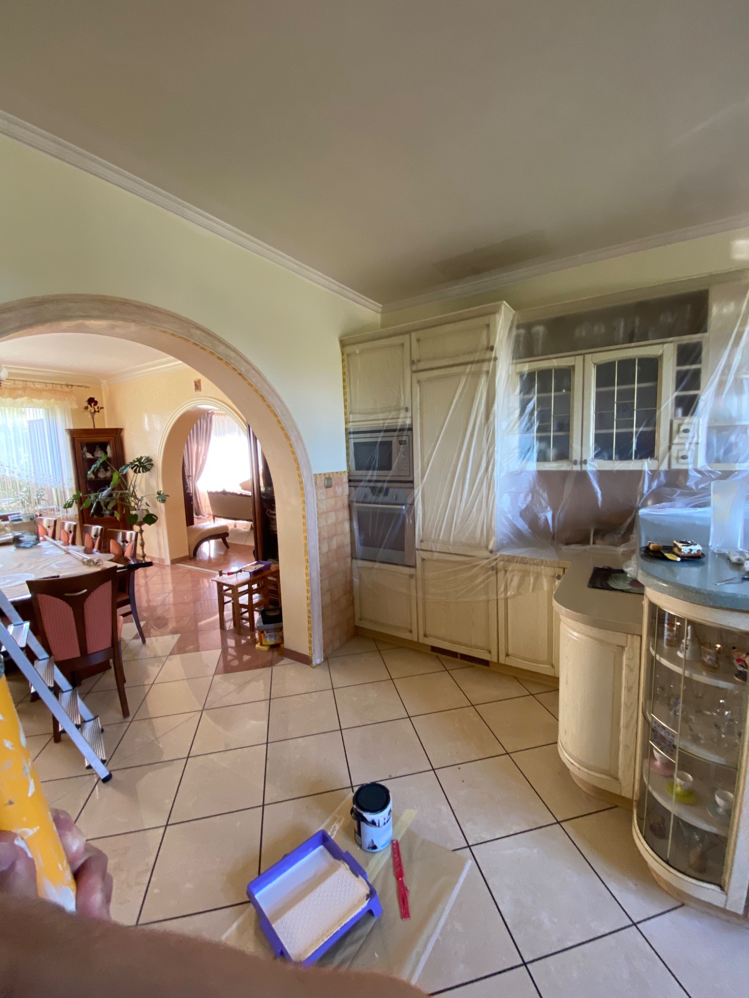 Przygotowania do malowania kuchni: zabezpieczone folią szafki, kuweta z wałkiem, puszka farby i drabina w tle łukowatego przejścia do jadalni.