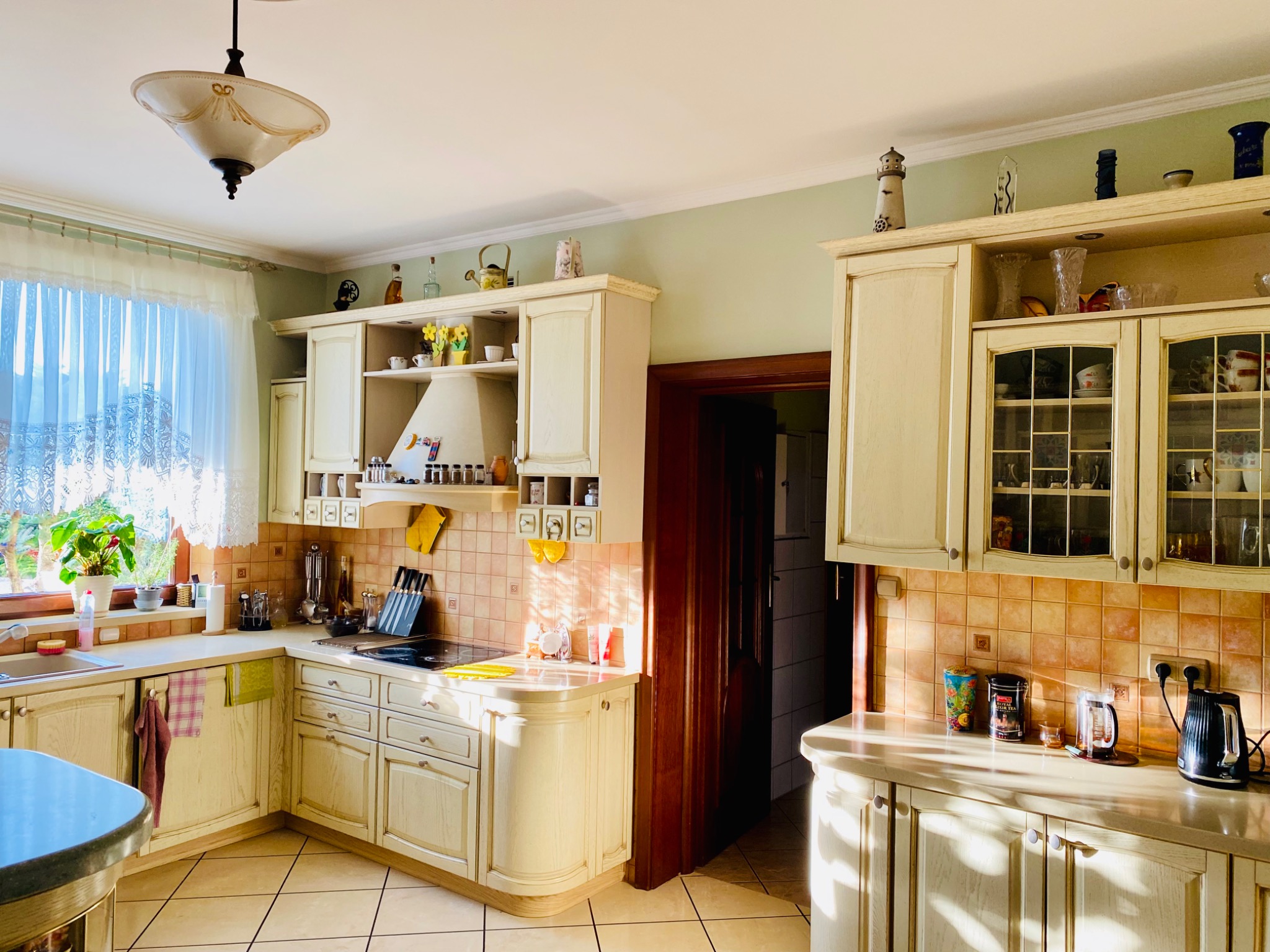 Jasna kuchnia z kremowymi szafkami w stylu rustykalnym, z dekoracyjnymi przedmiotami na półkach i mozaiką na ścianie, oświetlona naturalnym światłem wpadającym przez okno.