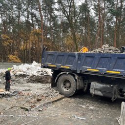 Rem-Ekspert Oleh Yefremov - Ciężarówka z gruzem po rozbiórce na tle lasu. Pracownik w kasku nadzoruje załadunek. Widoczny fragment wyburzonego muru i resztki materiałów budowlanych.