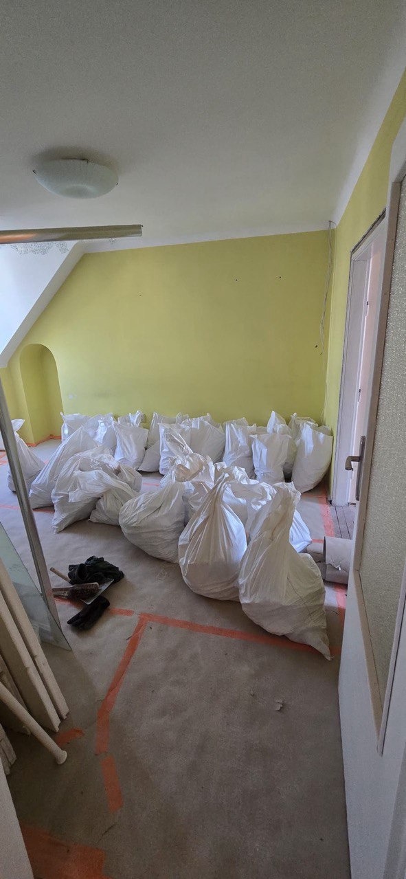 Pokój po remoncie: żółte ściany, podłoga z pomarańczowymi liniami i worki z gruzem. Widoczne narzędzia i fragmenty elementów budowlanych.