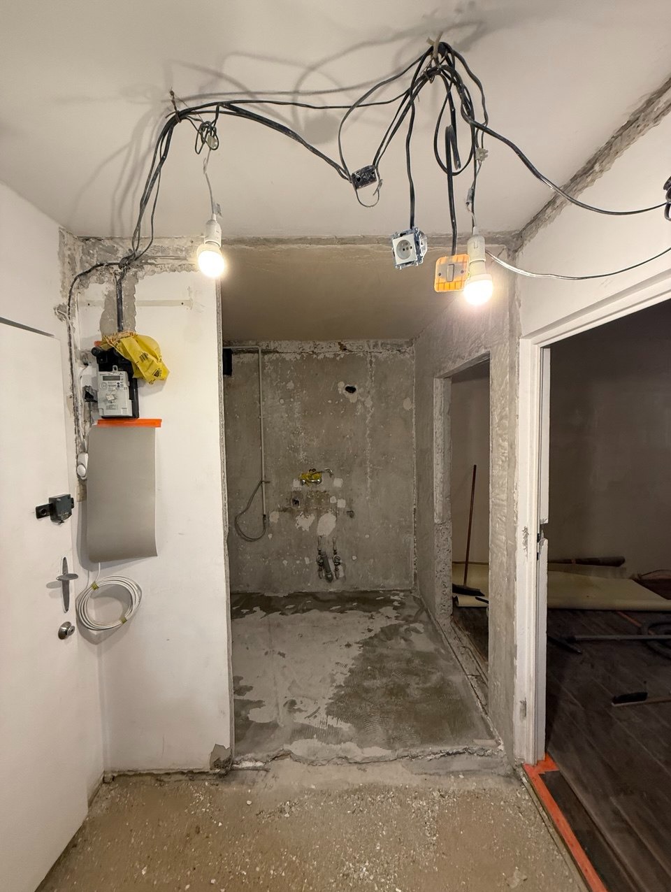 Wnętrze w trakcie remontu: surowe ściany, prowizoryczne oświetlenie z wiszącymi kablami, widoczne ślady po instalacjach. Podłoga częściowo przygotowana pod wylewkę.