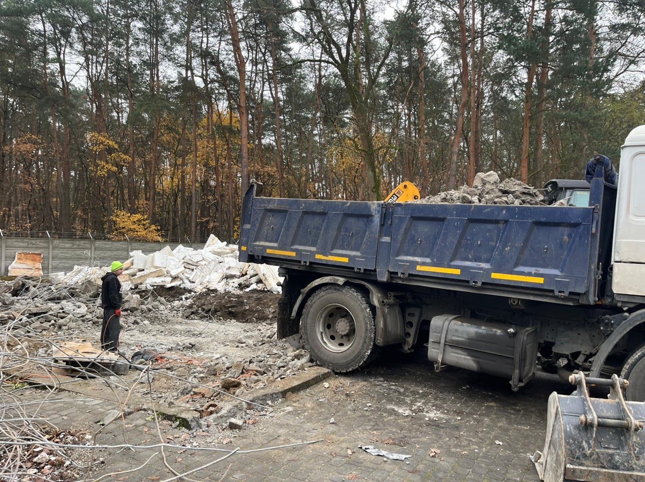 Ciężarówka z gruzem po rozbiórce na tle lasu. Pracownik w kasku nadzoruje załadunek. Widoczny fragment wyburzonego muru i resztki materiałów budowlanych.