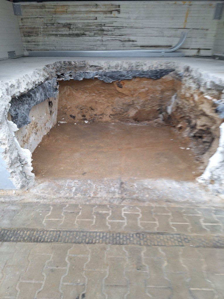 Wyburzona podłoga z odsłoniętą ziemią, widoczne fragmenty gruzu i izolacji. Prace przygotowawcze do remontu, z bliska, z perspektywy chodnika.