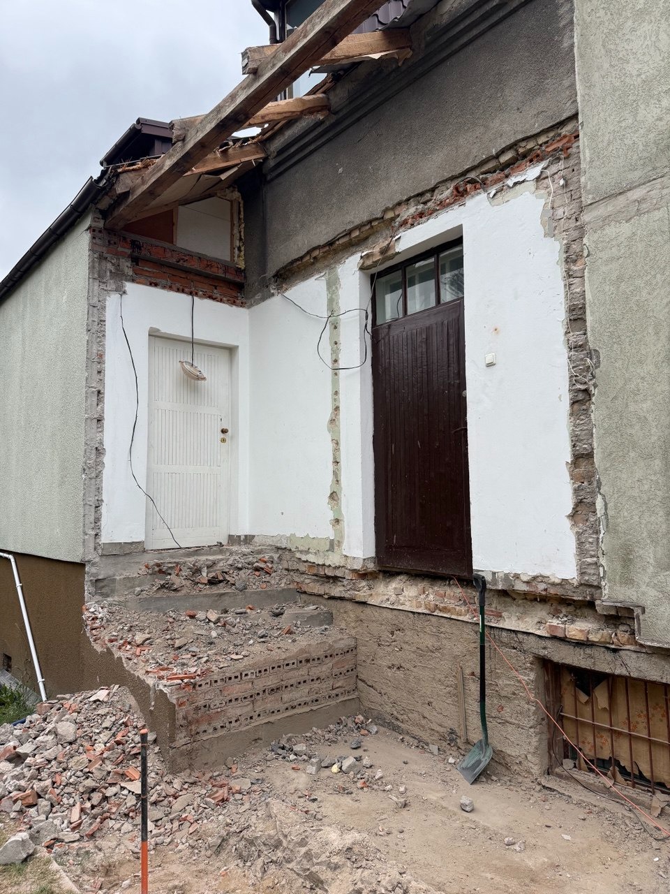 Częściowo rozebrany budynek z odsłoniętą konstrukcją dachu, gruzem na schodach i łopatą opartą o ścianę. Widoczne ślady po wyburzeniach.