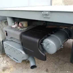 Elementy podwozia samochodu dostawczego po renowacji: zbiornik powietrza i osłona pomalowane na czarno i szaro. Widoczne narzędzia malarskie i pędzle.