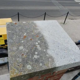 Ceglany słupek z betonową płytą: połowa wyczyszczona z mchu i zabrudzeń, druga pokryta nalotem. Widoczny żółty agregat czyszczący i element ulicy w tle.