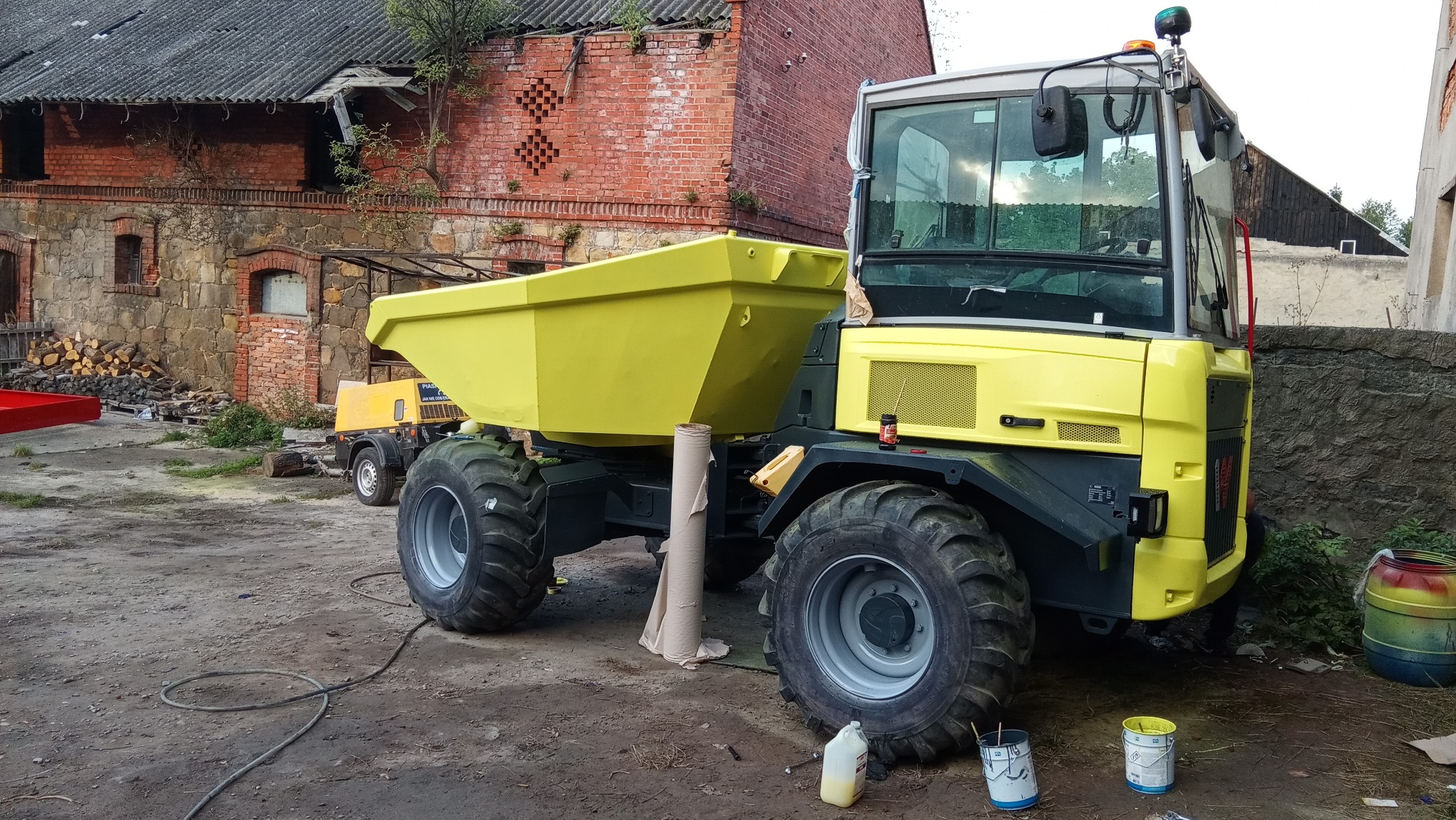 Żółta wywrotka po renowacji, z widocznymi śladami farby i narzędziami, na tle starego budynku. Nietypowy kąt i oświetlenie podkreślają detale.