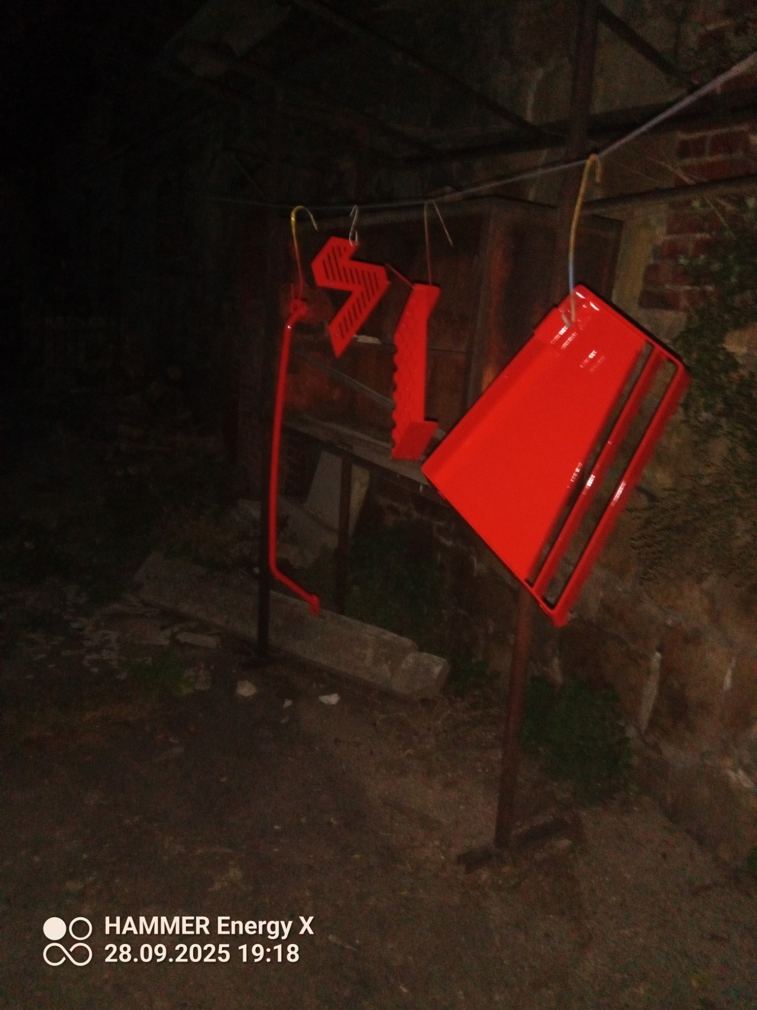 Czerwone, metalowe elementy konstrukcyjne wiszą na haku po obróbce. Widoczne ślady farby i niedoskonałości, ciemne otoczenie.