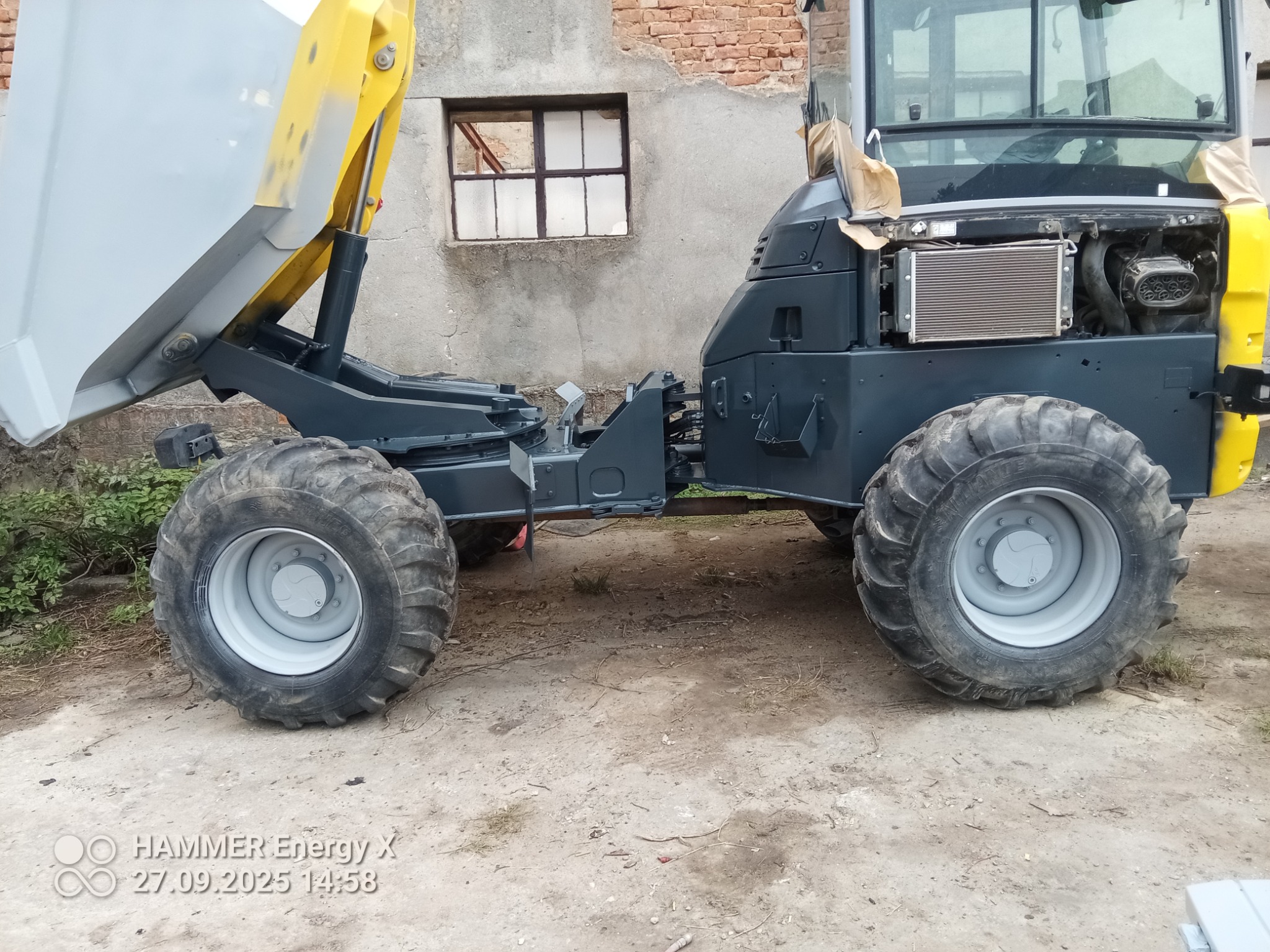 Wyremontowana wywrotka z podniesioną skrzynią, pomalowana na szaro i żółto, stoi na zewnątrz. W tle budynek z cegły z oknem.