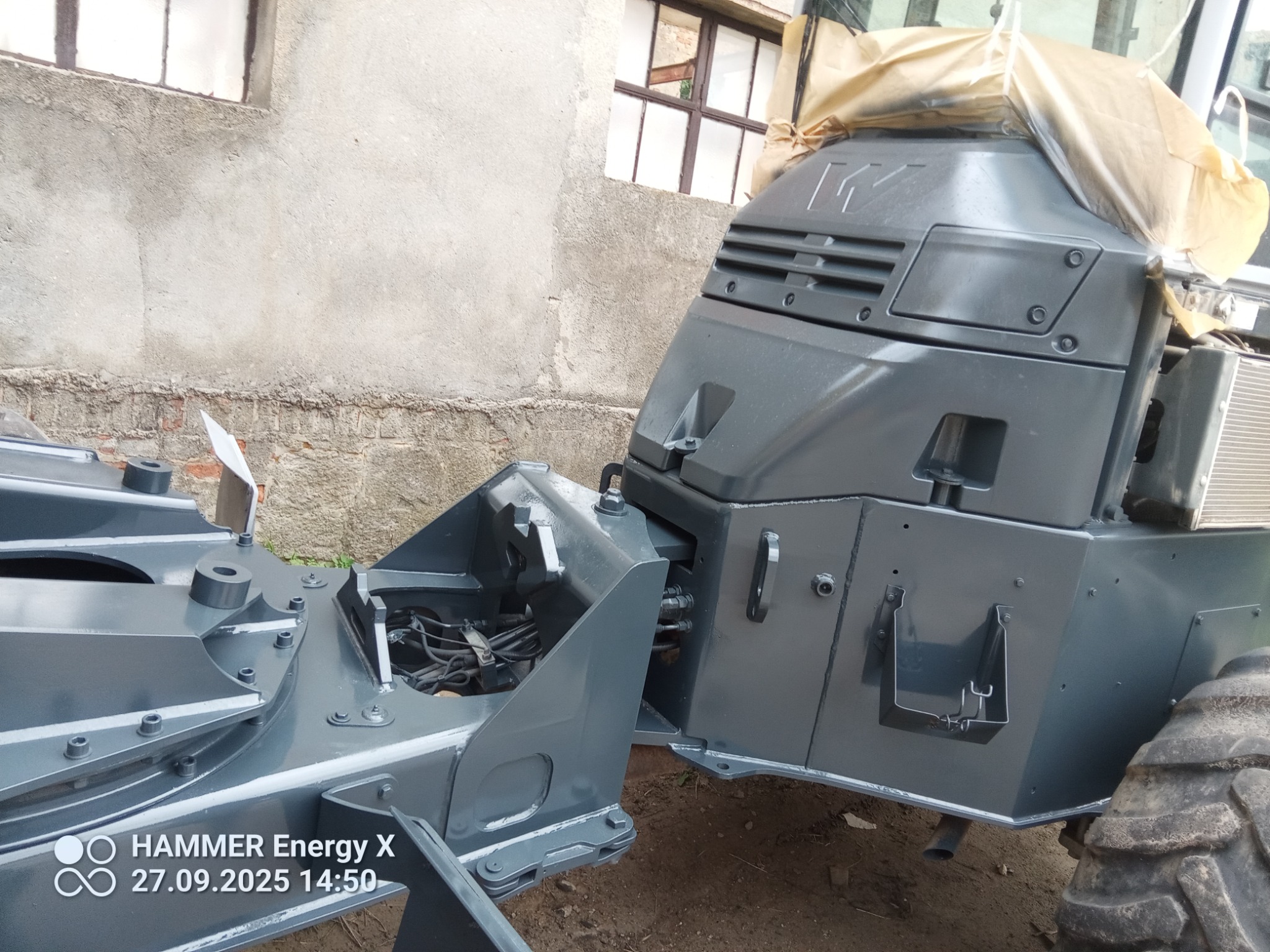 Szare elementy konstrukcyjne pojazdu budowlanego po renowacji. Widoczne łączenia, śruby i fragment opony. Część elementów osłonięta folią malarską.