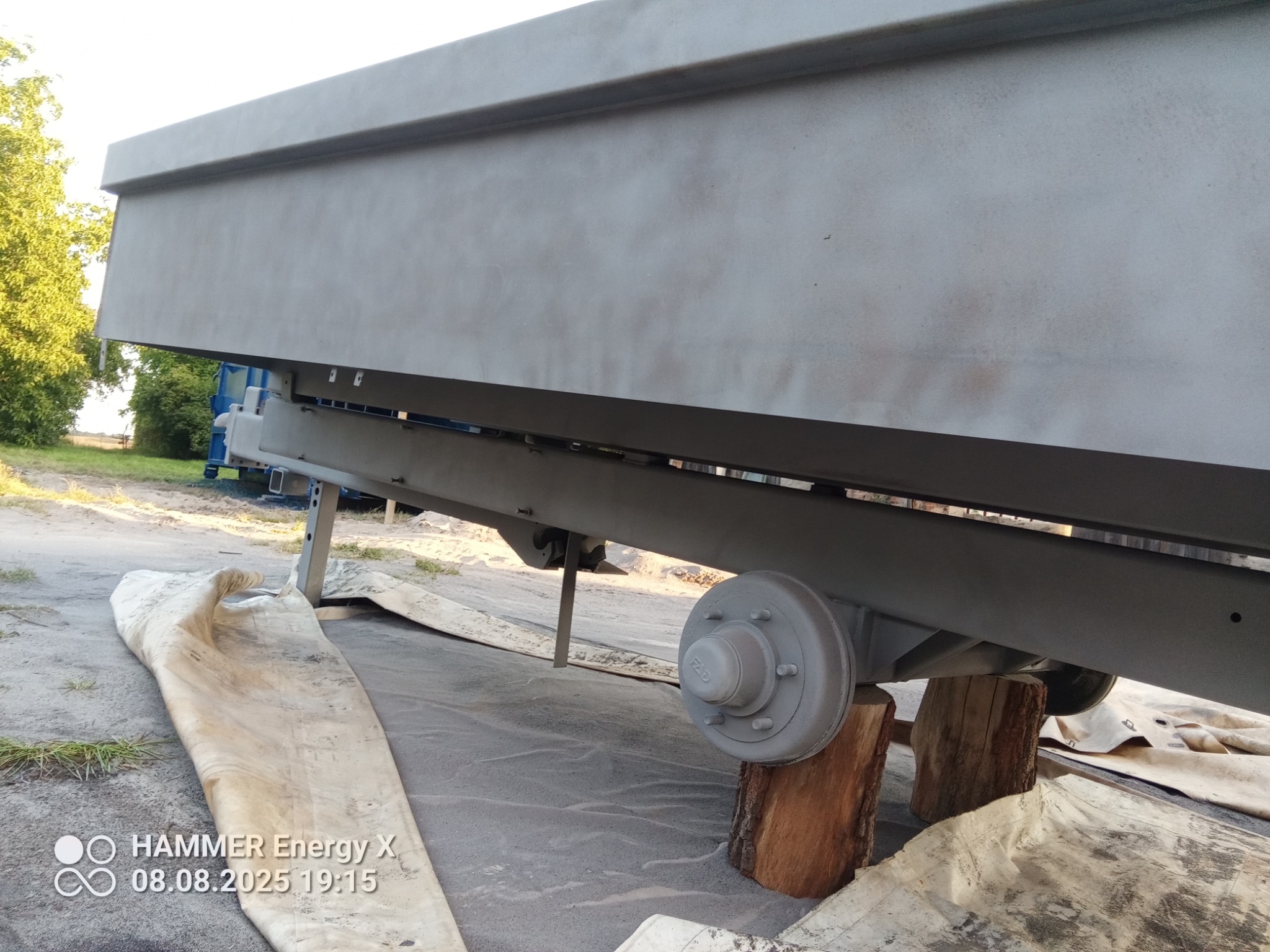 Przyczepa po renowacji, zabezpieczona plandeką, wsparta na drewnianych klockach. Szary, matowy kolor.