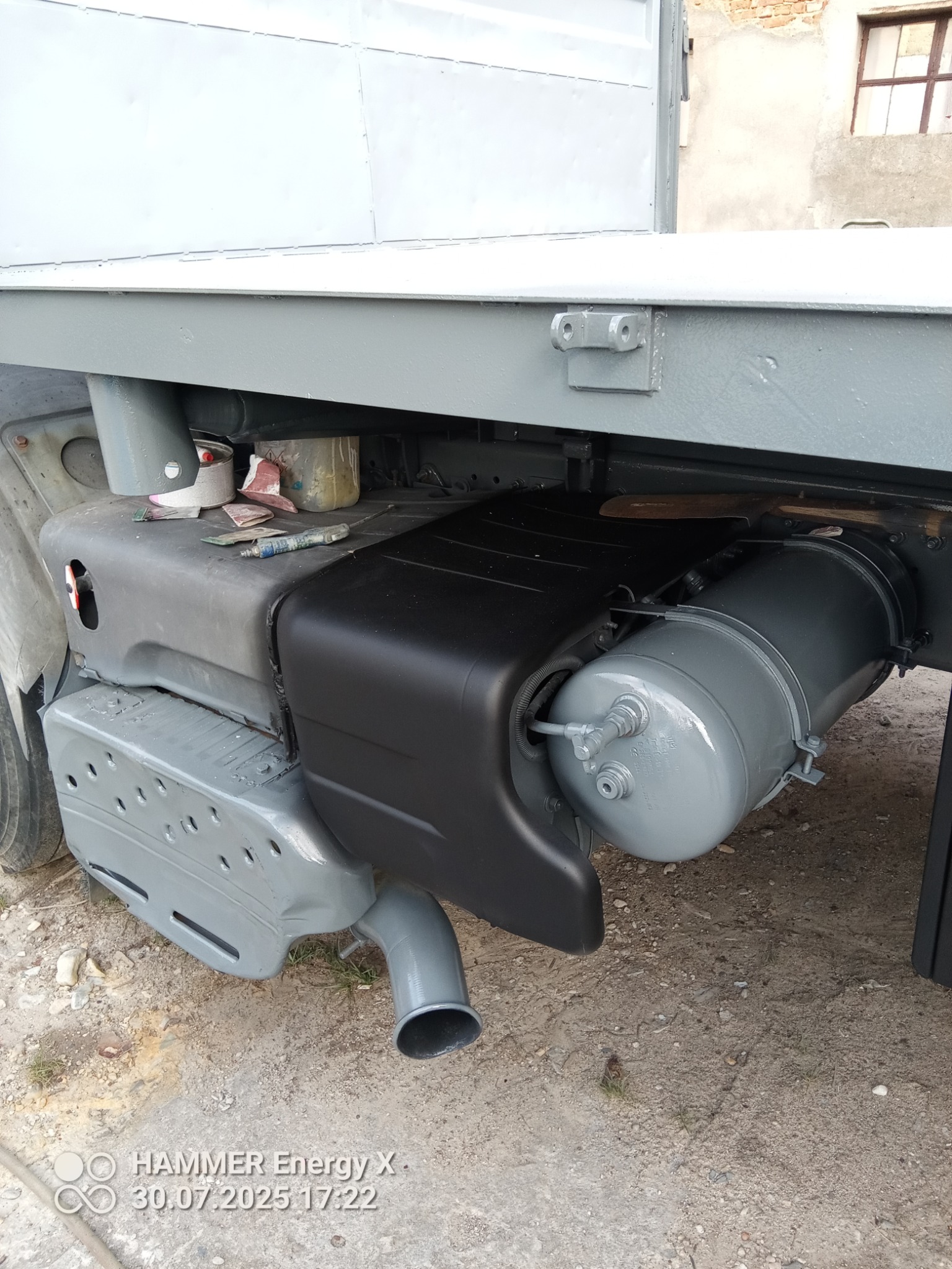 Elementy podwozia samochodu dostawczego po renowacji: zbiornik powietrza i osłona pomalowane na czarno i szaro. Widoczne narzędzia malarskie i pędzle.