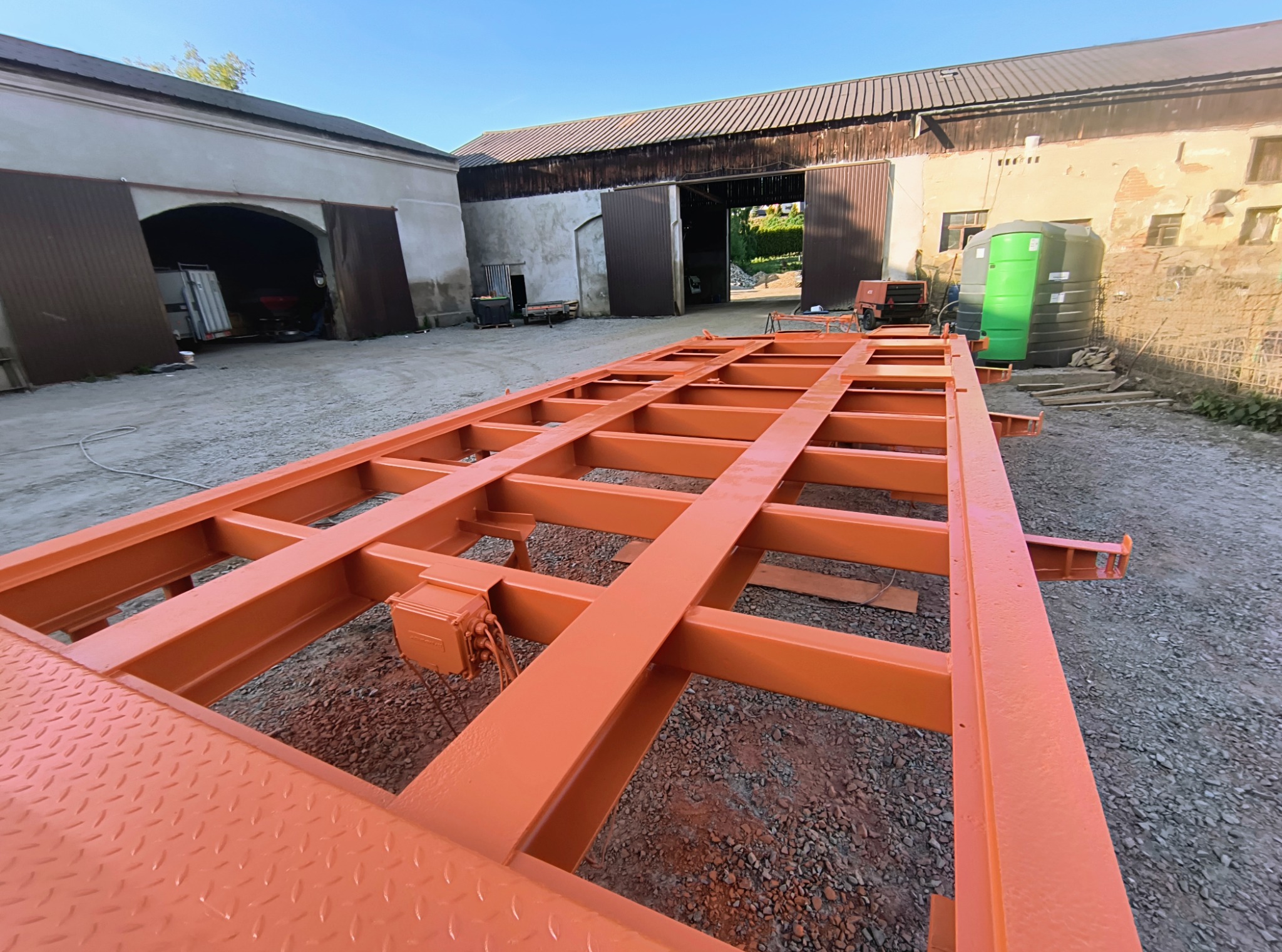 Pomarańczowa, pomalowana konstrukcja stalowa ramy przyczepy na żwirowym podłożu, na tle budynków gospodarczych w słoneczny dzień.