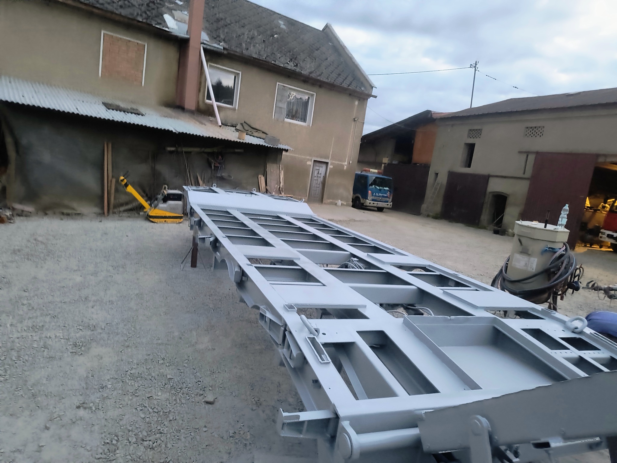 Szkielet przyczepy po obróbce, świeżo pokryty szarym lakierem. W tle budynek gospodarczy i pojazd. Zastosowano perspektywę z dołu.