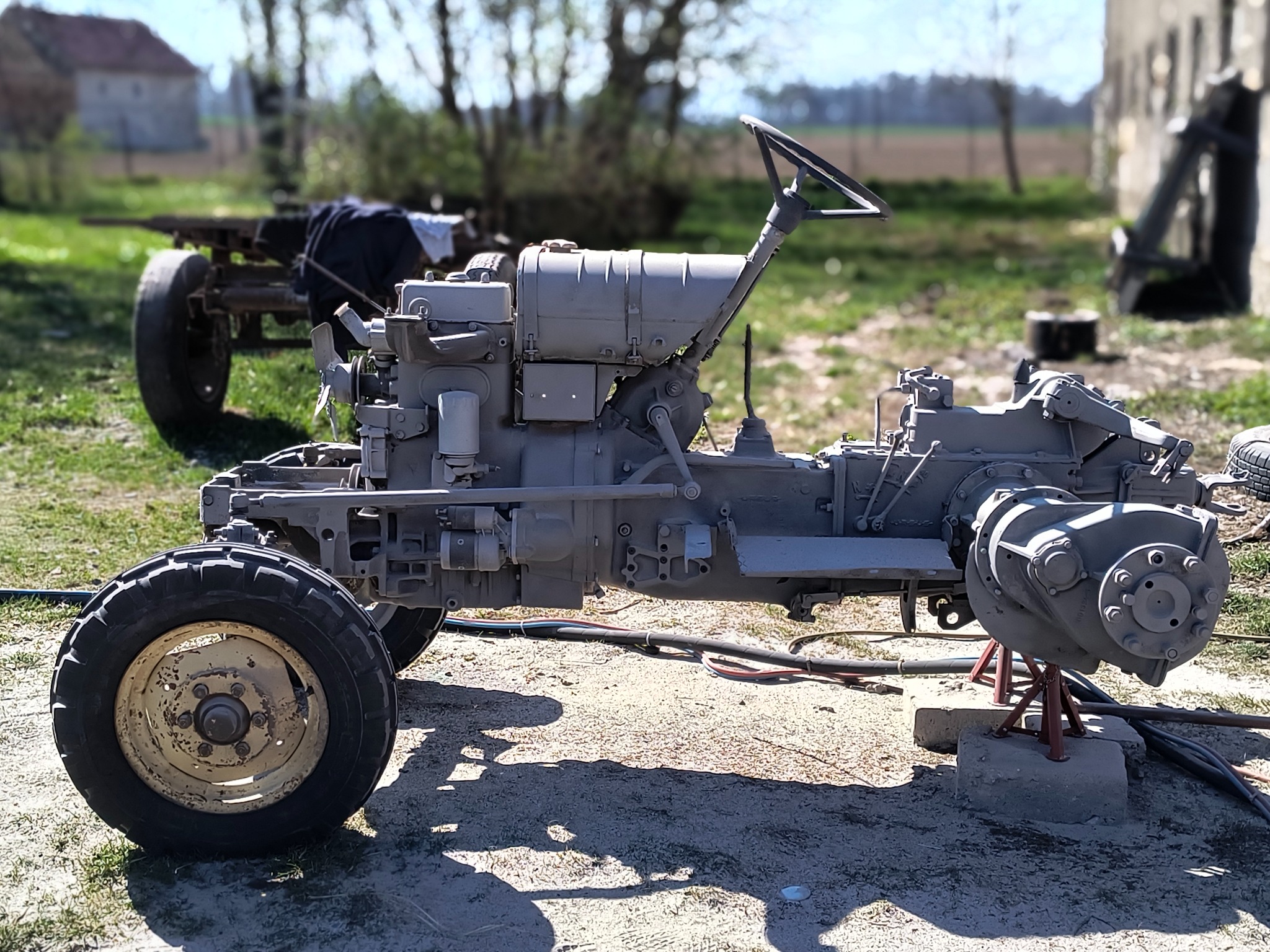 Odrestaurowany traktor rolniczy, pomalowany na szaro, stoi na zewnątrz. Widoczne detale mechaniczne i opony. W tle budynek gospodarczy i zieleń.