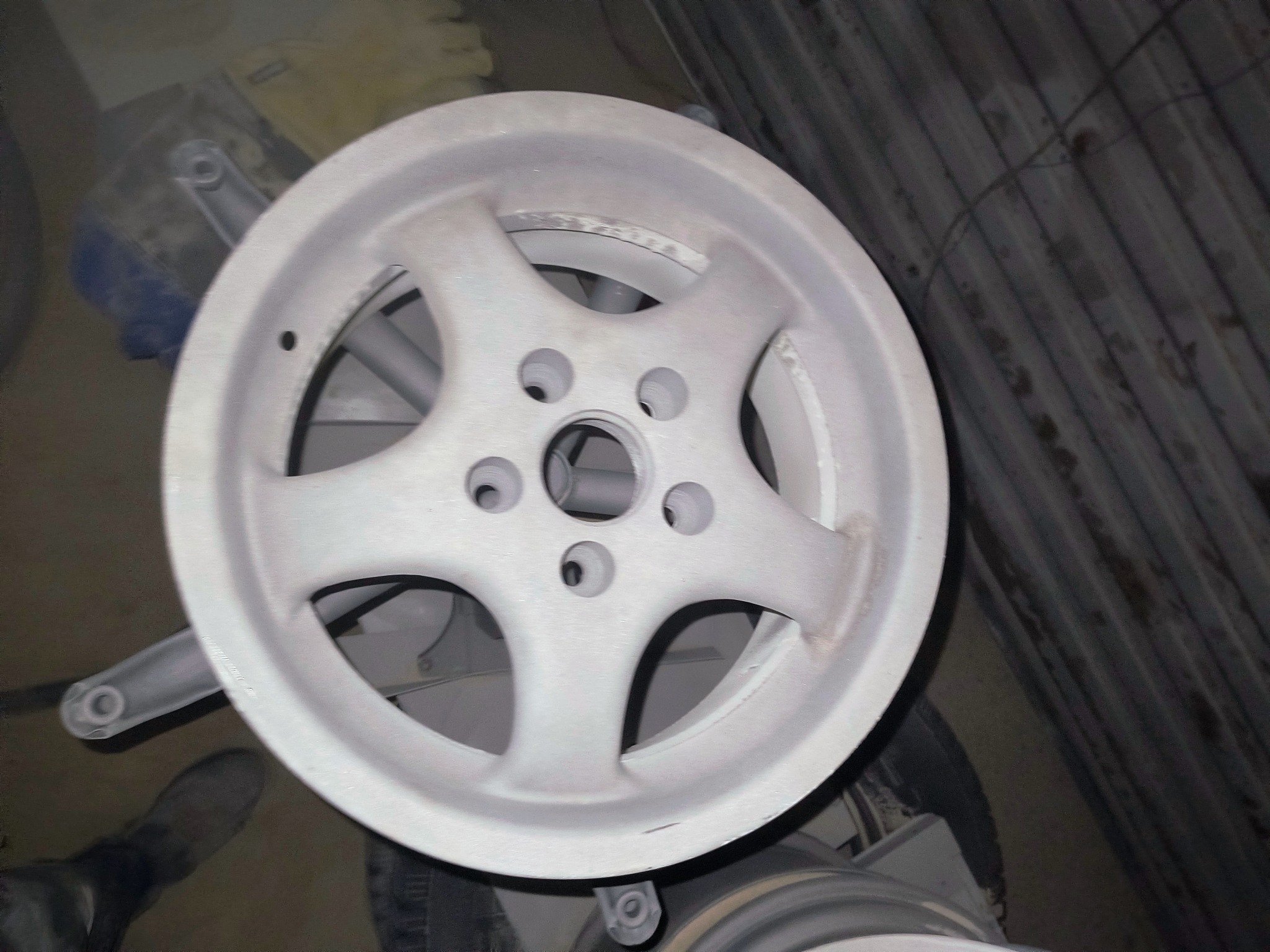 Szara felga aluminiowa z pięcioramiennym wzorem, leżąca na podłożu. Widoczne ślady po obróbce. Elementy zawieszenia w tle.