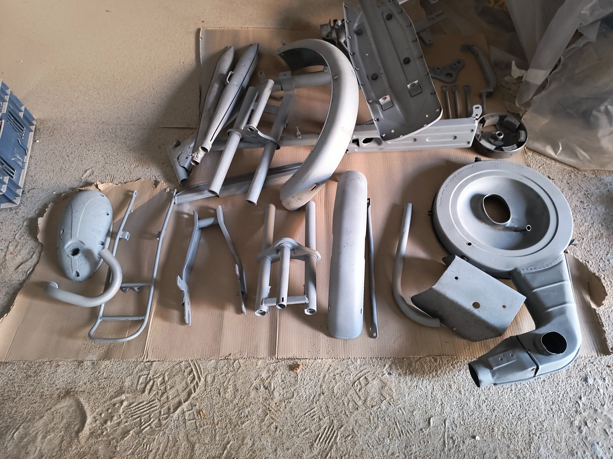 Elementy motocyklowe po piaskowaniu, przygotowane do malowania proszkowego. Widoczne błotniki, ramy i inne części w szarym kolorze na kartonie.