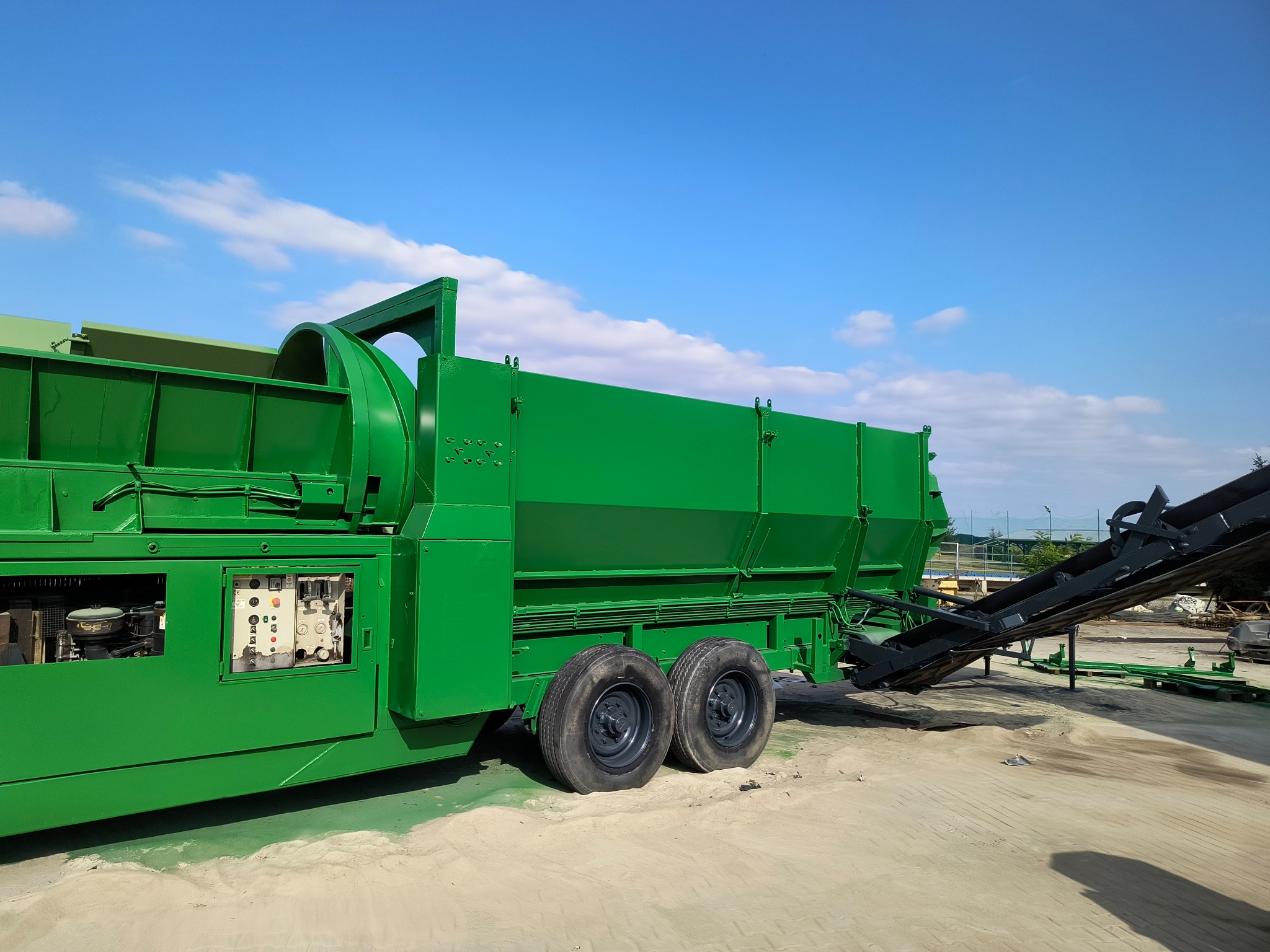 Zielona maszyna przemysłowa z podajnikiem na piasku, pod błękitnym niebem. Widoczna kontrolka i silnik. Świeżo pomalowana powierzchnia.