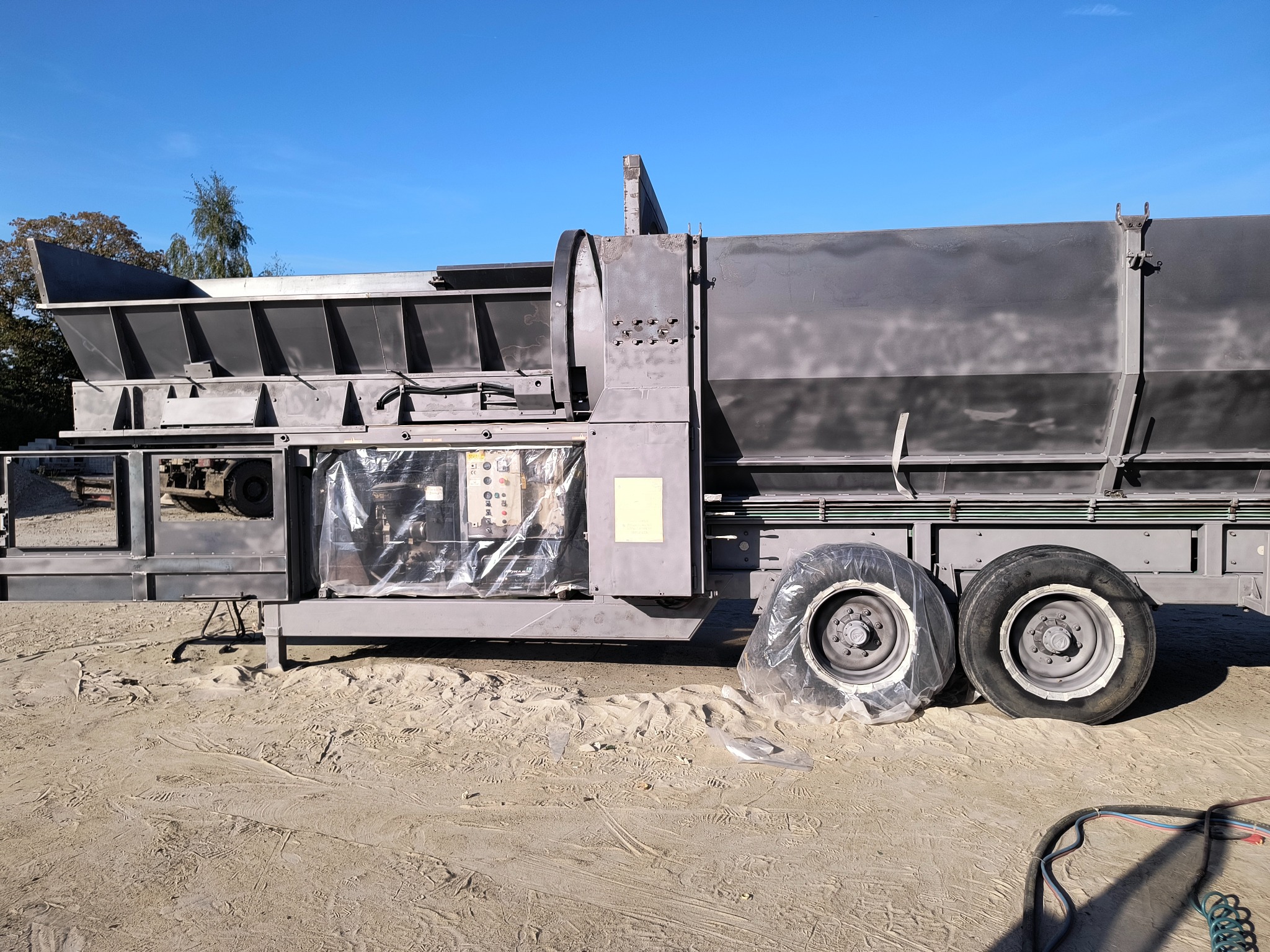 Duża, szara maszyna do obróbki powierzchni na piaszczystym terenie. Część maszyny osłonięta folią, widoczne koła i przewody.