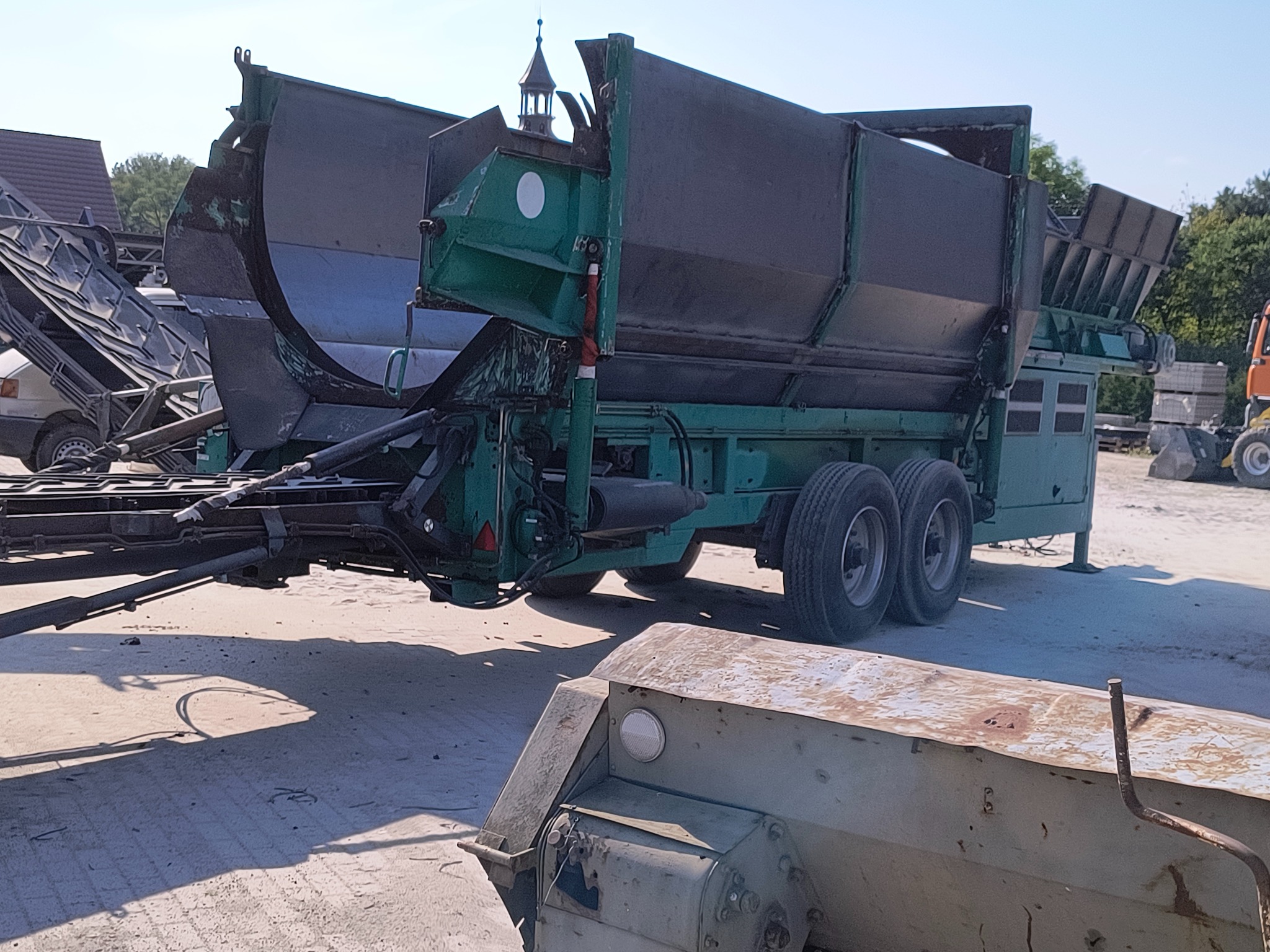 Zielona maszyna do obróbki strumieniowo-ściernej na podwoziu, z widocznymi śladami eksploatacji i rdzawymi plamami, na tle placu i zieleni.