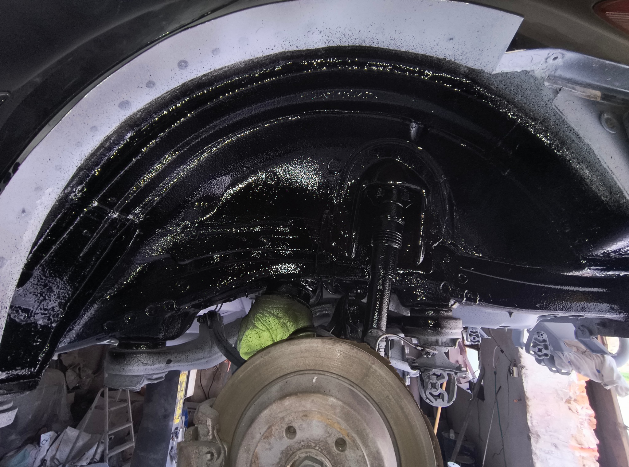 Wnętrze nadkola samochodu po zabezpieczeniu antykorozyjnym, pokryte czarną, błyszczącą powłoką. Widoczna tarcza hamulcowa i elementy zawieszenia.