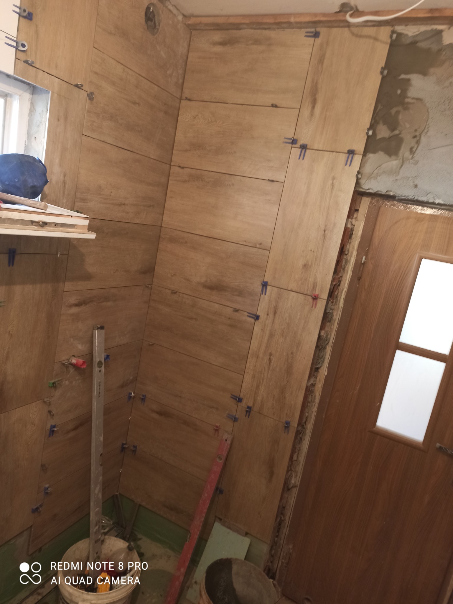Ściana łazienki w trakcie układania płytek imitujących drewno, z widocznymi krzyżykami dystansowymi, poziomica oparta o ścianę, oraz fragment ościeżnicy drzwi i surowego muru
