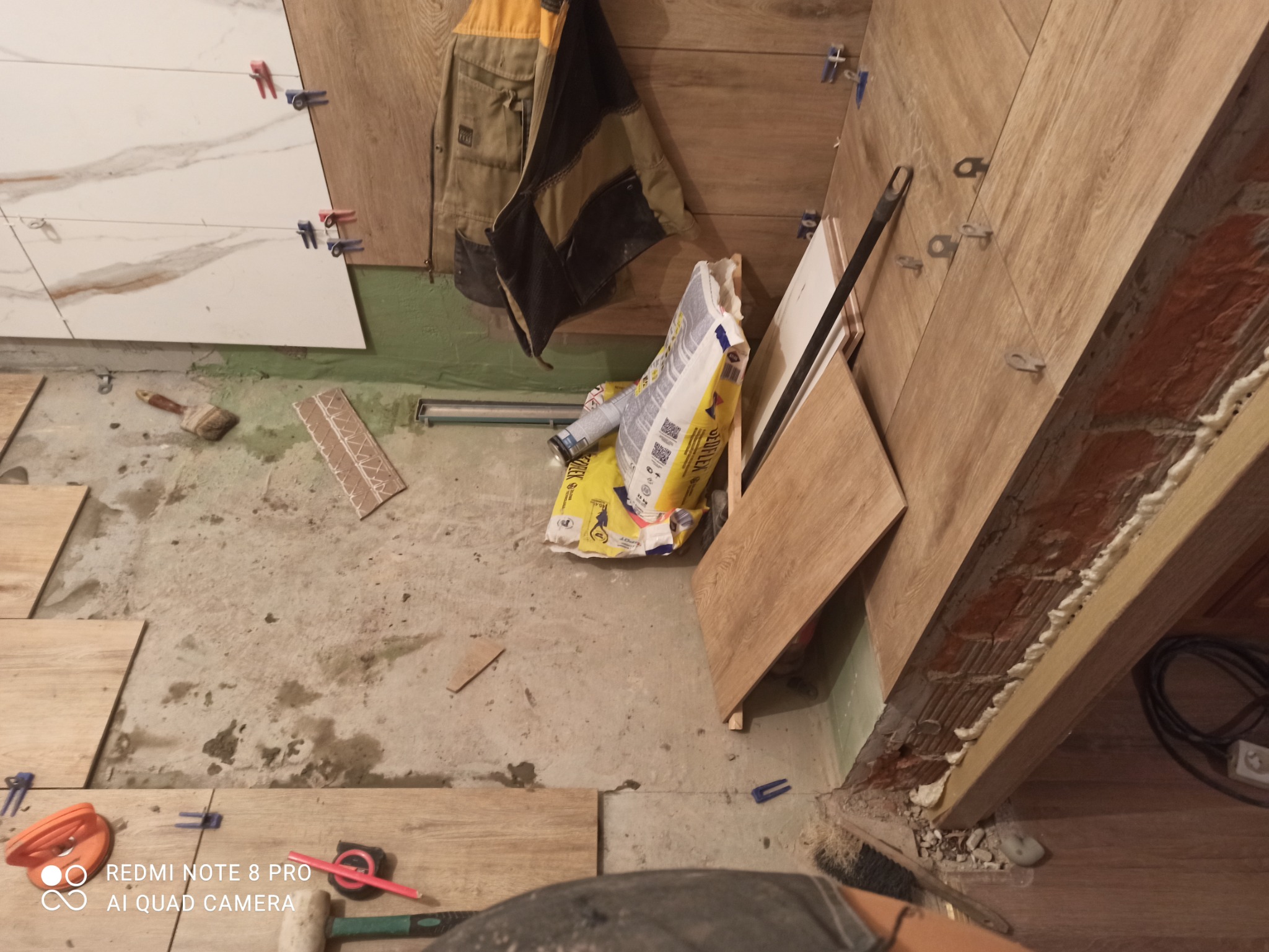 Układanie płytek imitujących drewno na ścianie, widoczne krzyżyki dystansowe, paczka kleju i robocza kurtka w tle.
