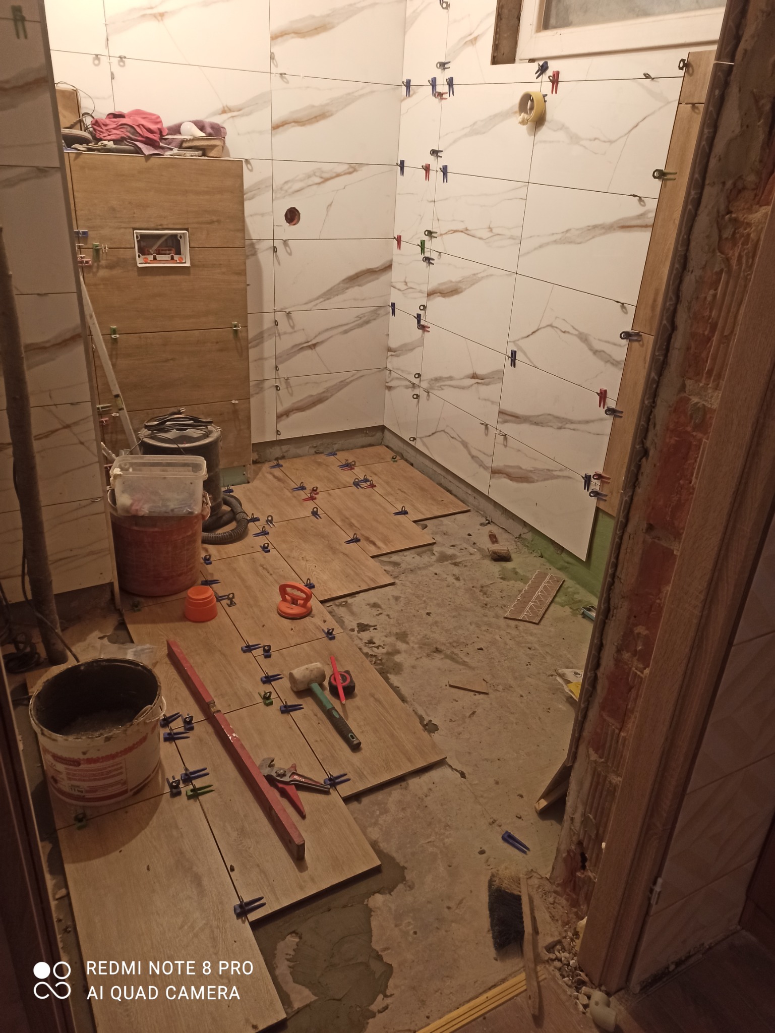 Układanie płytek imitujących drewno na podłodze i ścianach w łazience w trakcie remontu, widoczne narzędzia glazurnicze, system poziomowania płytek z klipsami i klinami, częściowo ułożone płytki...