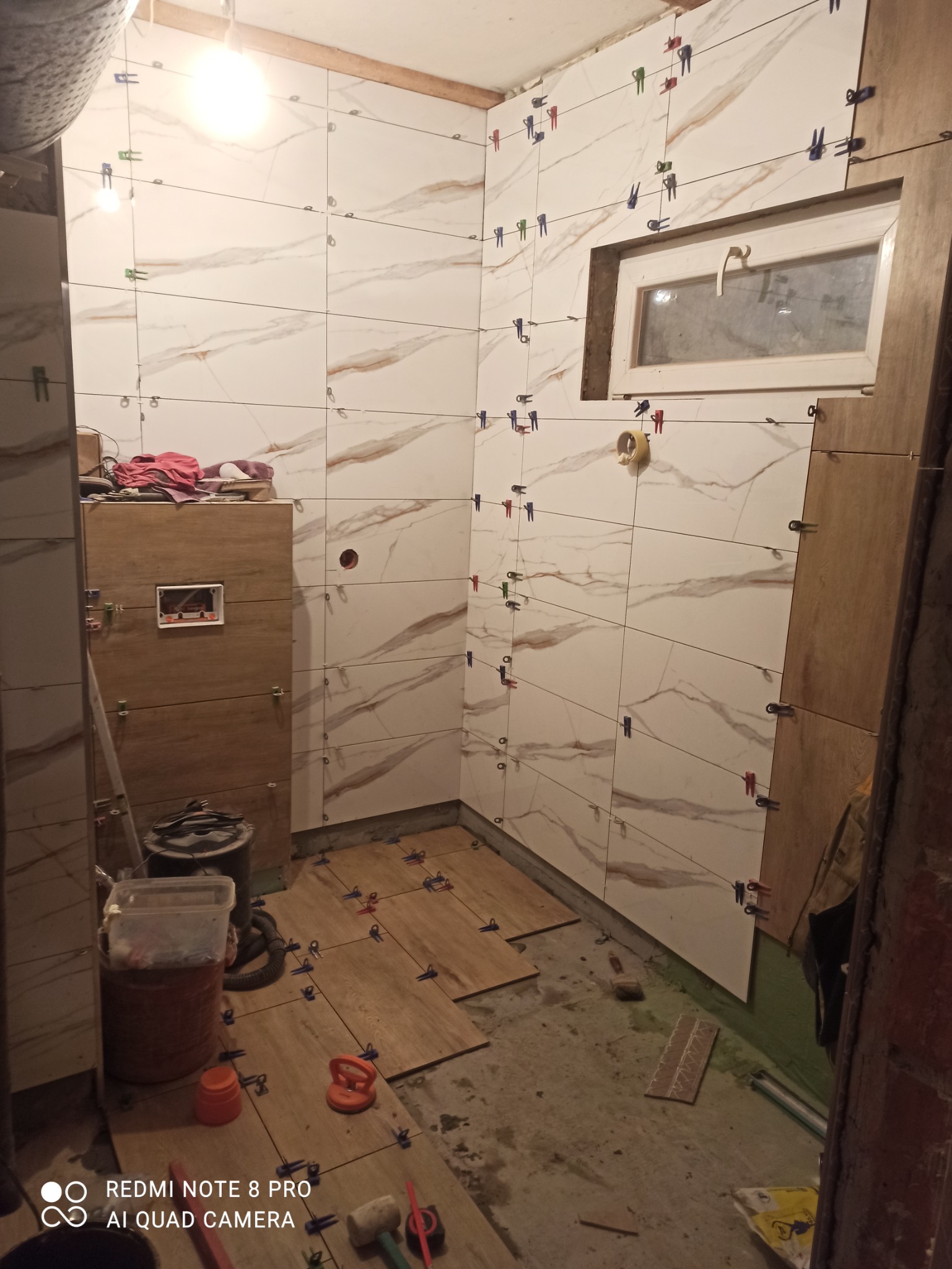 Wnętrze łazienki w trakcie układania płytek marmurkowych na ścianach i drewnopodobnych na podłodze, widoczne kolorowe klipsy dystansowe, narzędzia glazurnicze i okno.