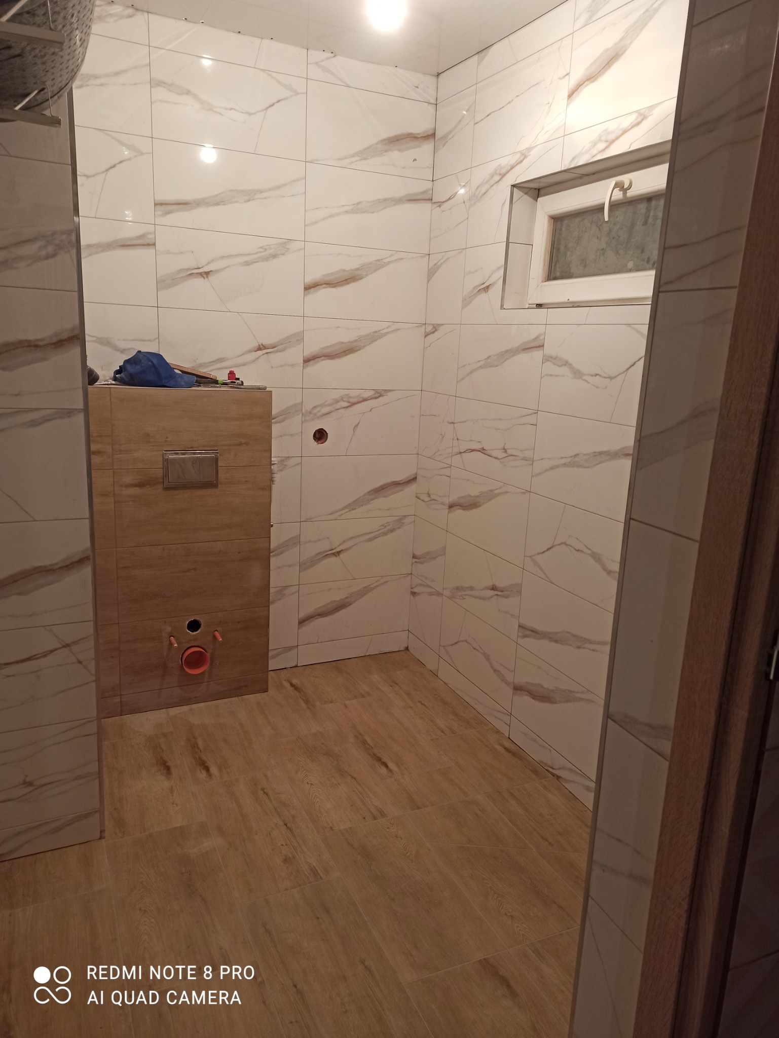 Wnętrze łazienki w trakcie wykańczania: położone płytki imitujące marmur na ścianach, zabudowa stelaża podtynkowego WC z drewnopodobną okładziną oraz podłoga wyłożona panelami winylowymi...