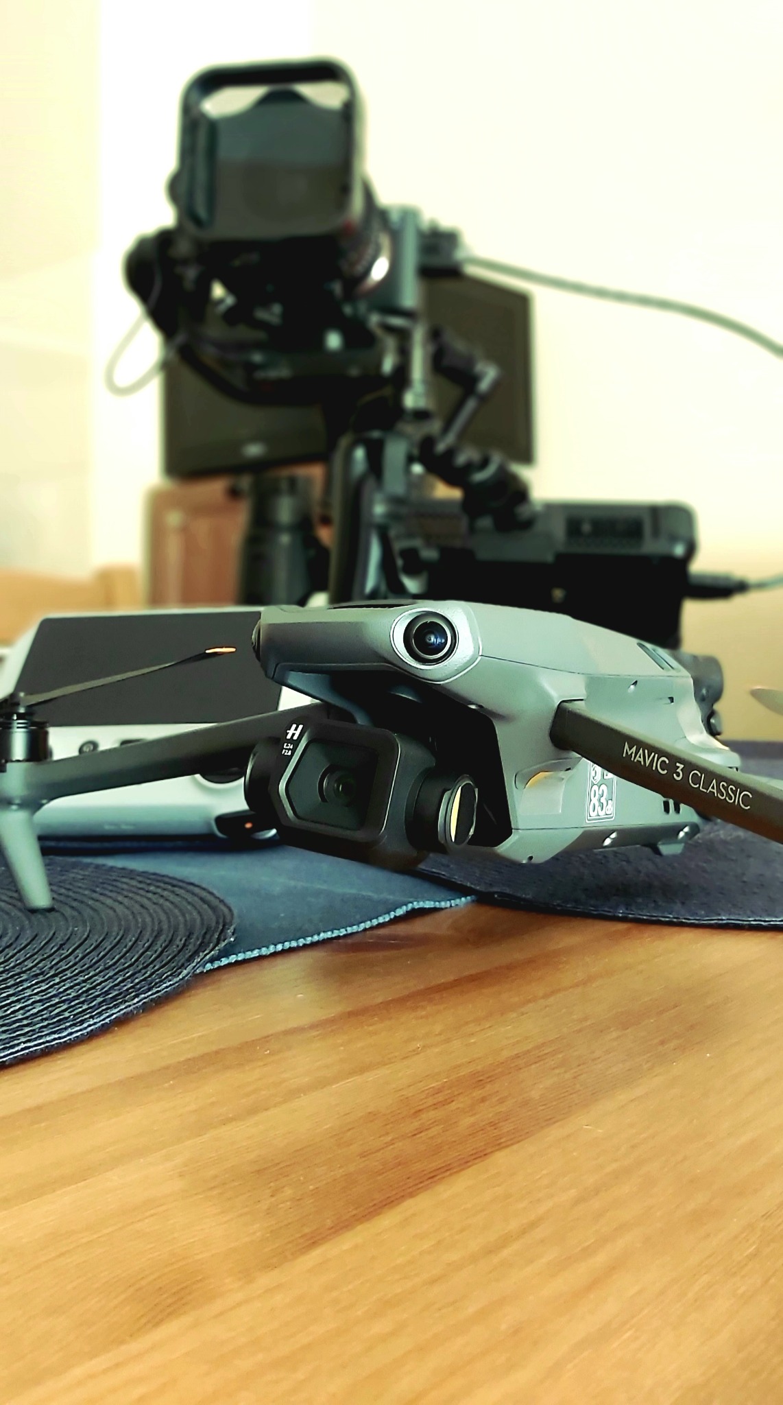Dron DJI Mavic 3 Classic na drewnianym stole, w tle rozmyta kamera na statywie i tablet, sugerujące przygotowania do filmowania.