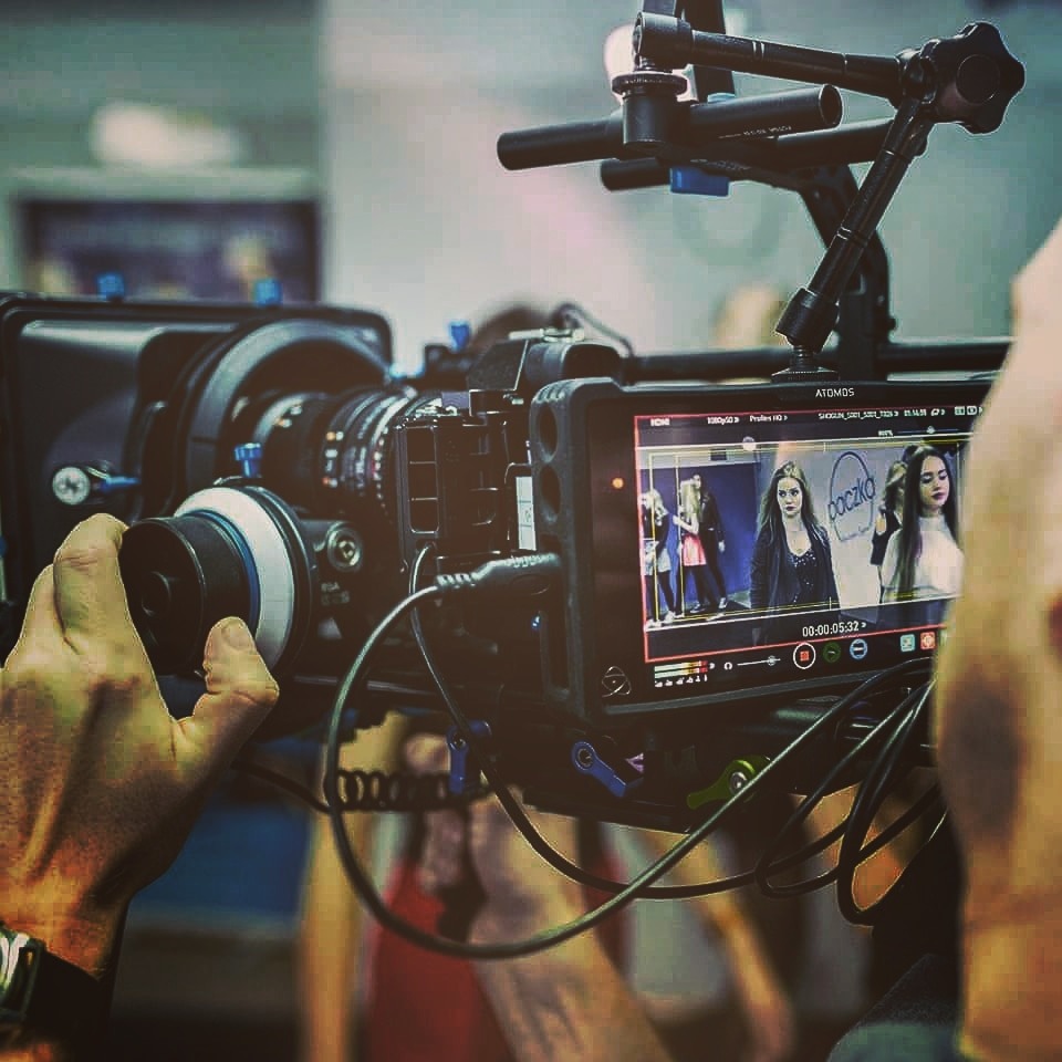 Operator filmowy obsługuje profesjonalną kamerę z podglądem nagrywanego materiału, na ekranie widoczne modelki podczas sesji.