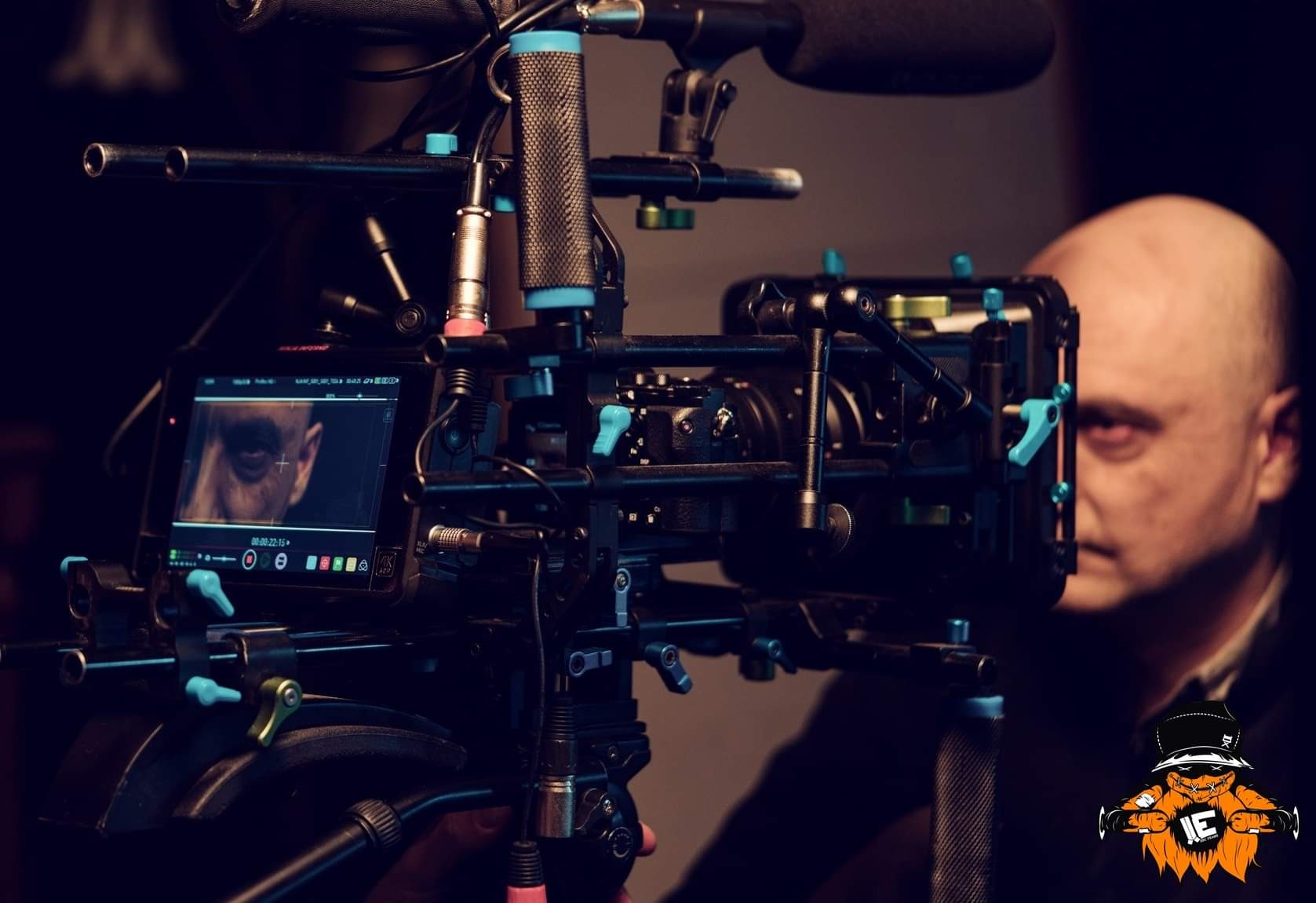 Profesjonalna kamera filmowa z podglądem na ekranie, na którym widoczny jest fragment twarzy filmowanej osoby. W tle widoczny zarys głowy mężczyzny.